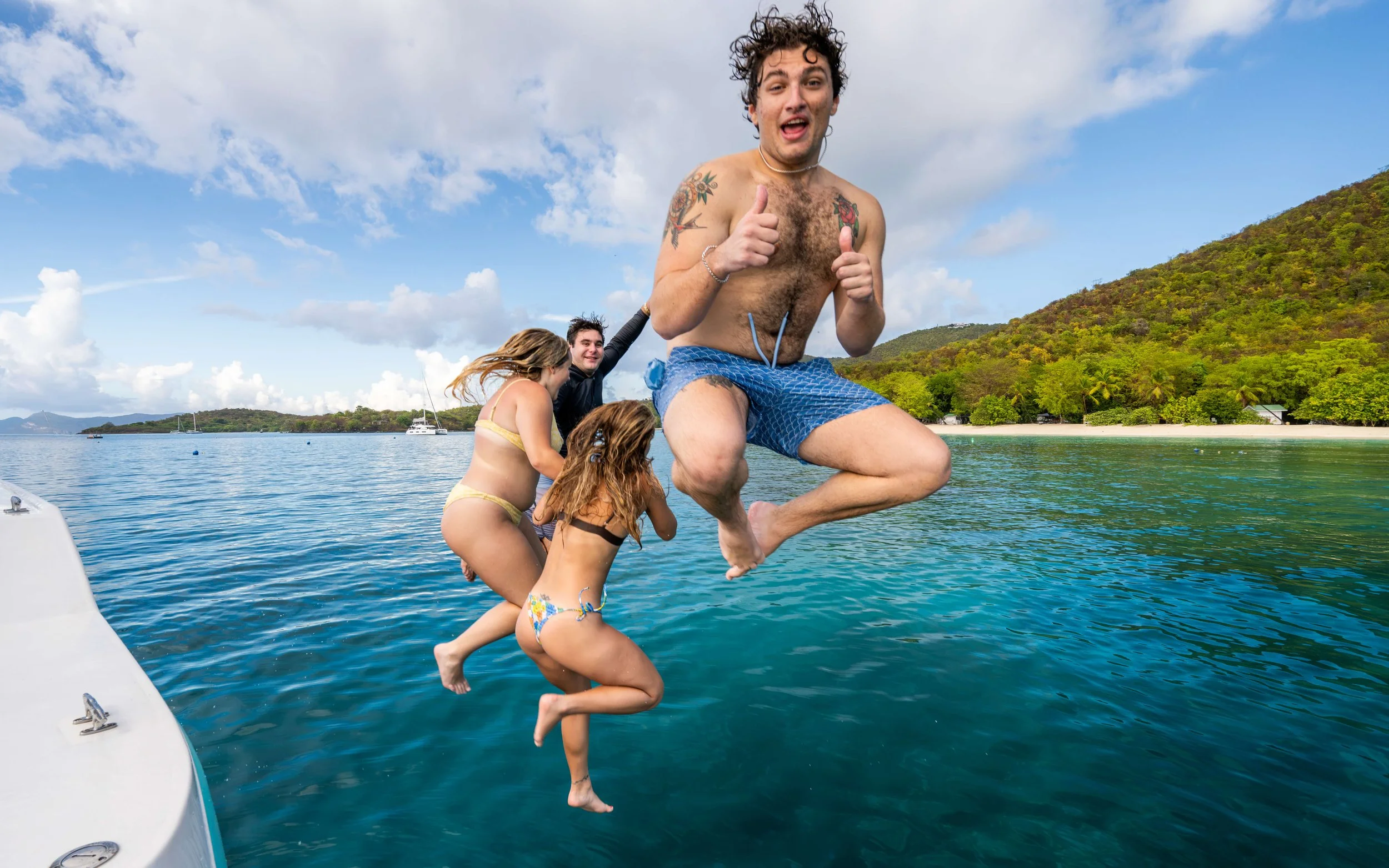 A group of four people on a boat, with one man jumping into the water, in a tropical location with green hills and blue sky.