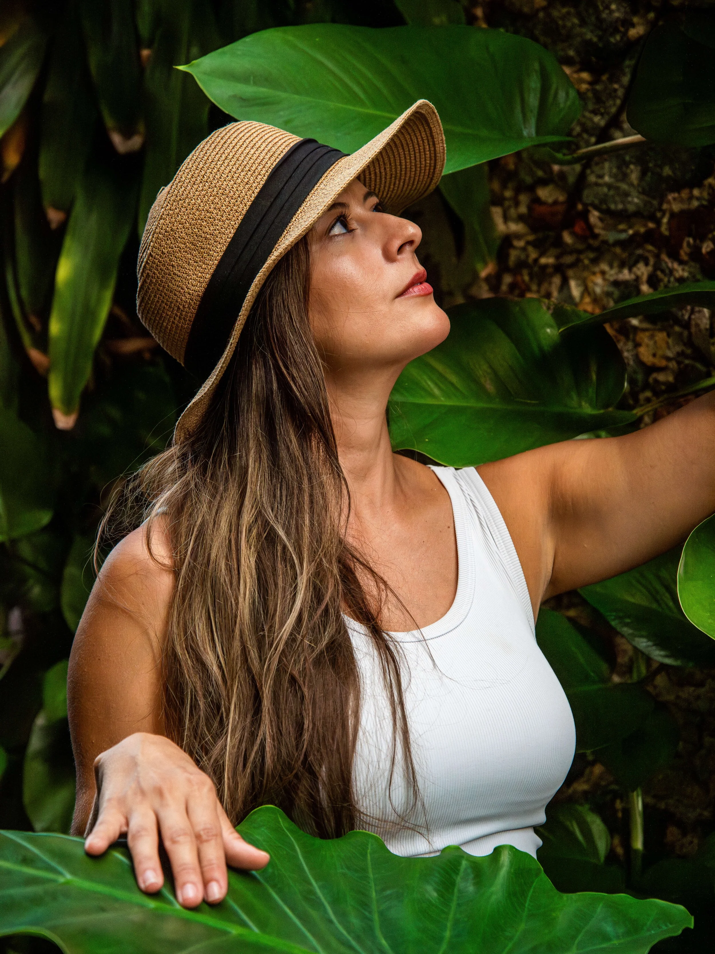 A woman with long brown hair wearing a wide-brimmed straw hat and a white sleeveless top, surrounded by large green tropical leaves.