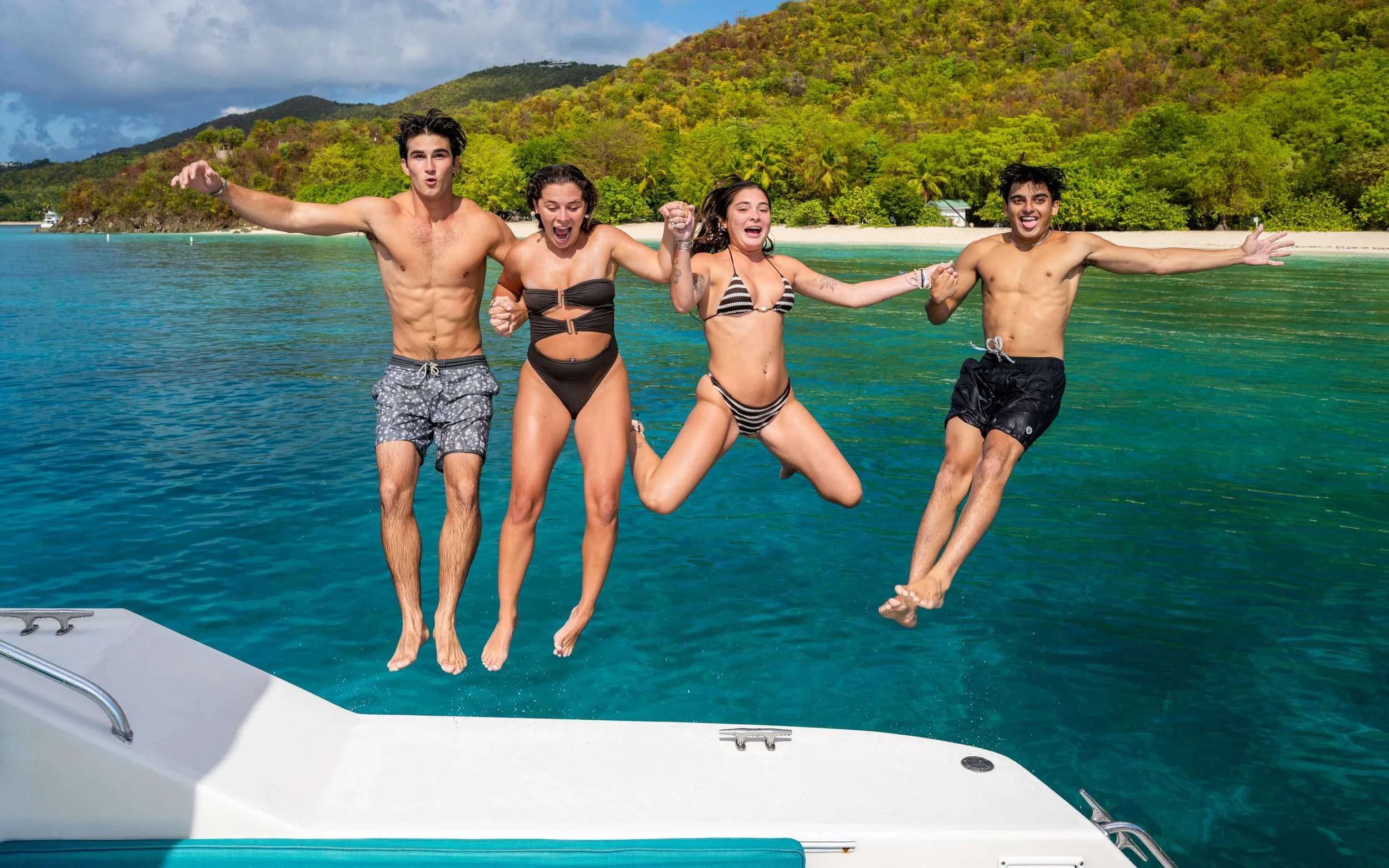 Four young people are jumping off a boat into clear blue water near a tropical island with lush green hills in the background. They are wearing swimsuits and appear to be enjoying a day on the water.