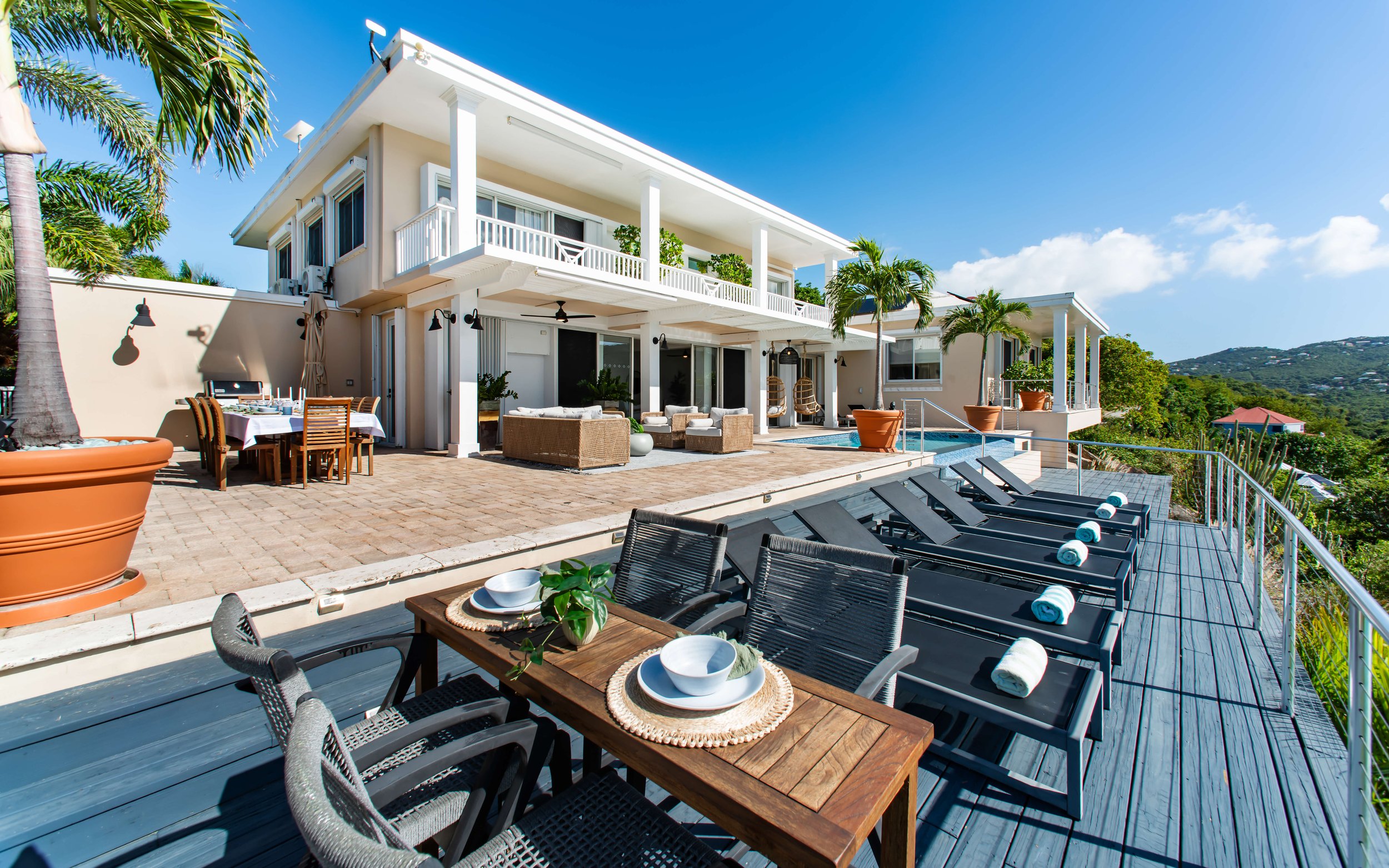 Luxury house with a large terrace featuring outdoor dining, seating areas, a swimming pool, and lounge chairs on a hillside with a mountain view under a clear blue sky.