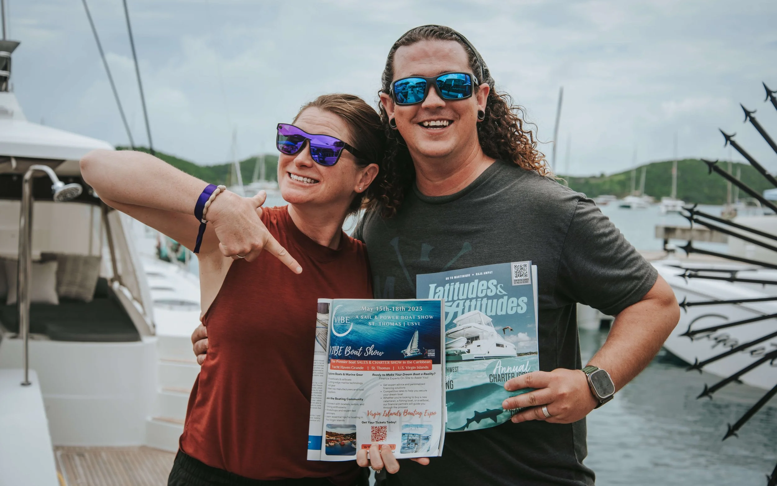 Two smiling people holding boating event flyers, wearing sunglasses, standing on a dock with boats in the background.