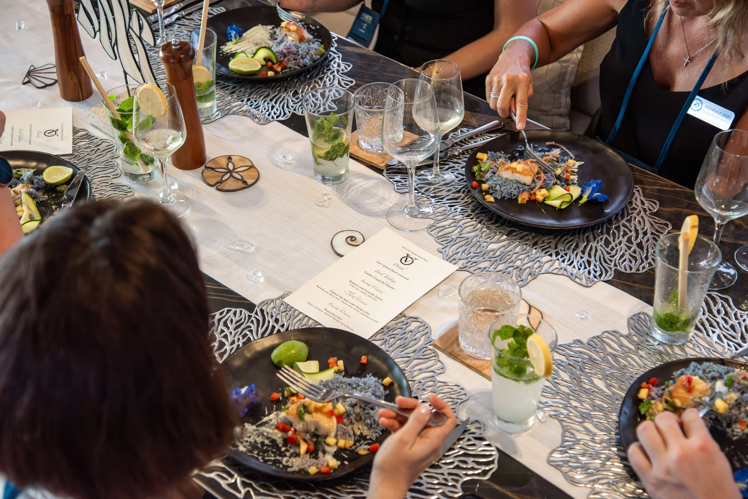 A group of people sitting at a dining table, enjoying a meal with plates of colorful food, glasses of beverages, and decorative table settings.
