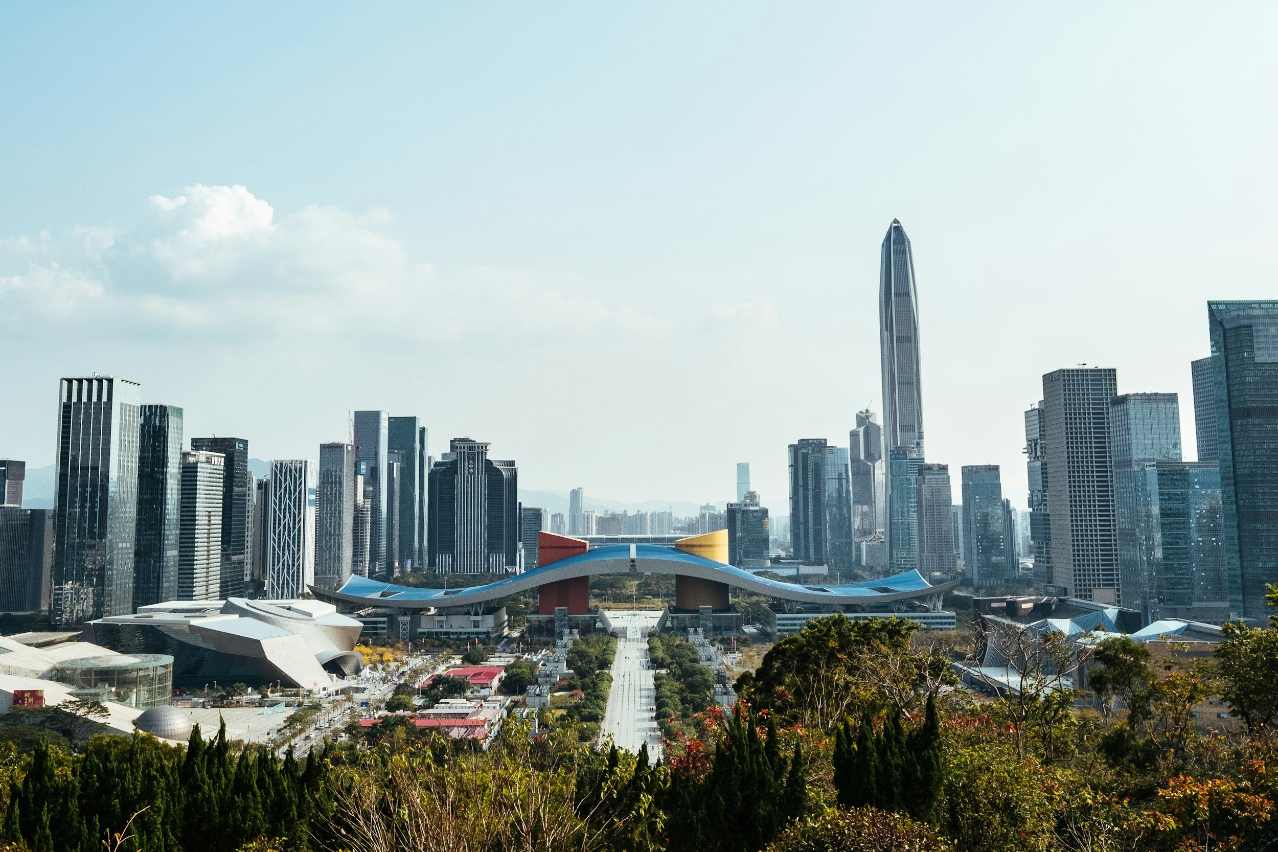 City skyline with tall skyscrapers and modern architecture, with greenery in the foreground and a blue sky with clouds.