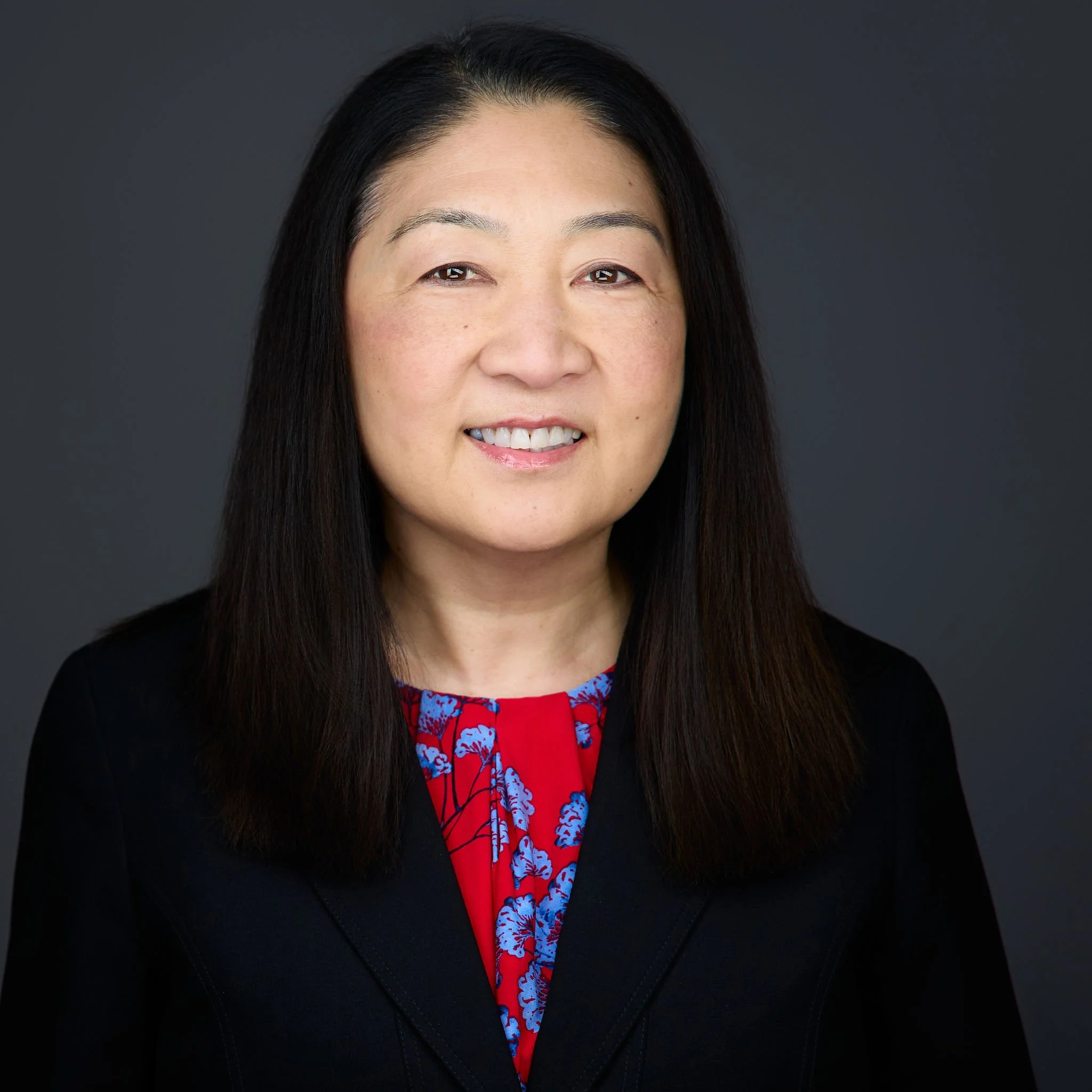 Professional headshot of Marina Saito with black hair wearing a black blazer and a red and blue patterned blouse, smiling against a gray background.