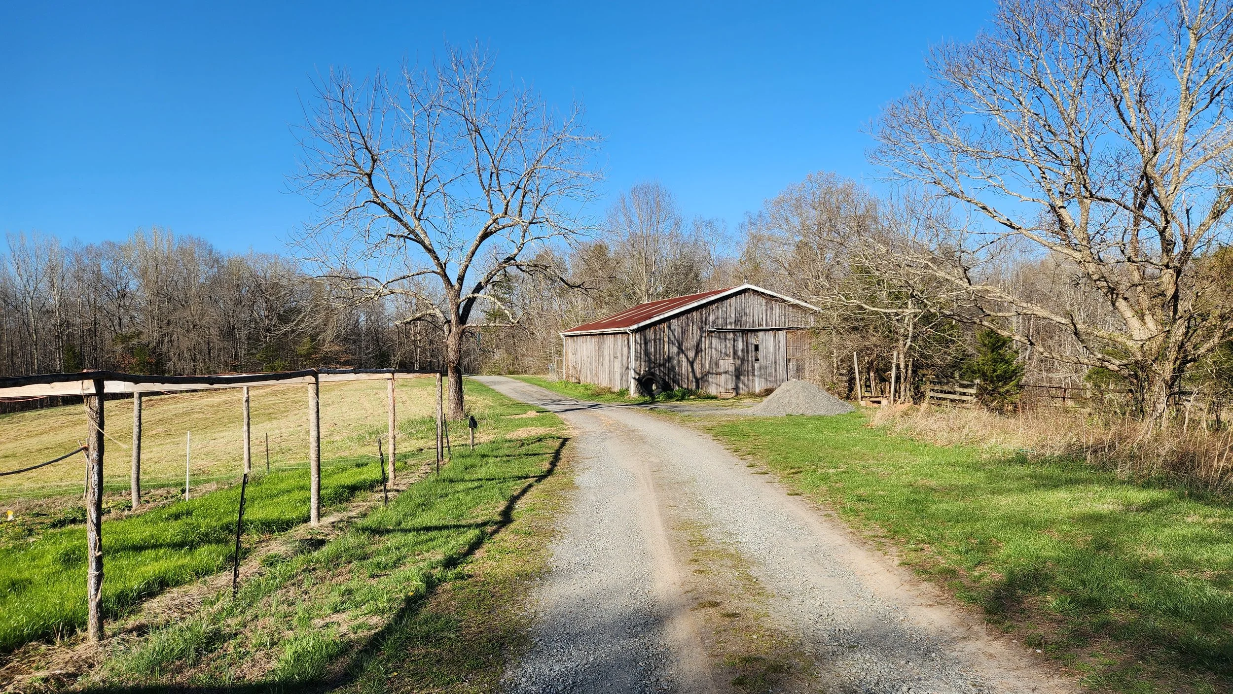 A gravel driveway leading toward an old wooden barn with a rusted metal roof, surrounded by leafless trees and a wooden fence, under a clear blue sky.
