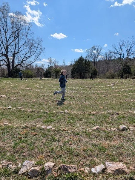 Child running across a grassy field with scattered rocks, trees, and a blue sky with clouds.
