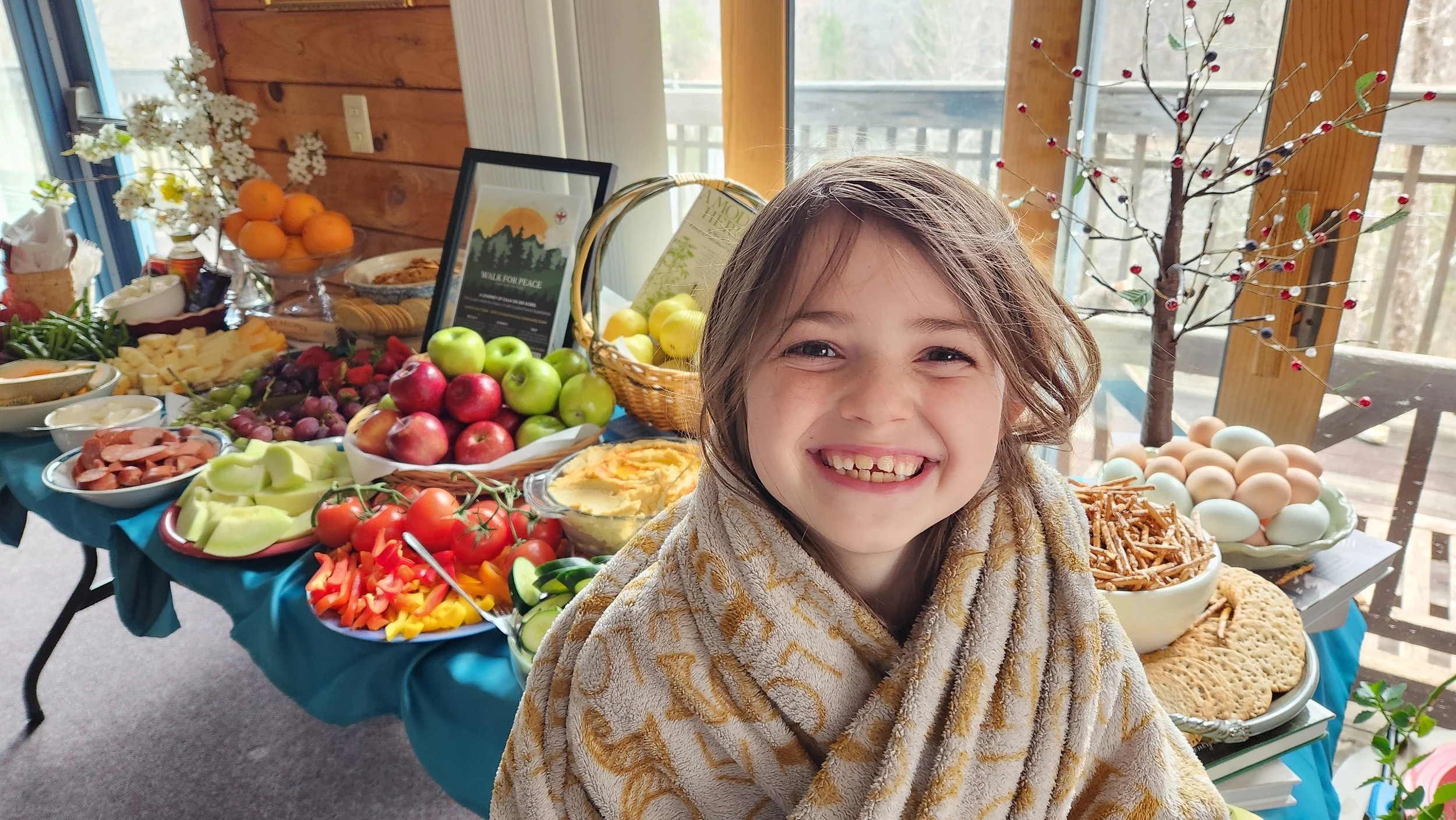 A young girl smiling in front of a table filled with various breakfast foods, including fruits, eggs, and baked goods, in a bright room with wooden walls and large windows.
