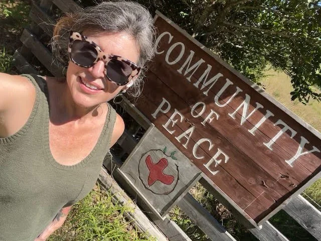 A woman taking a selfie outdoors next to a wooden sign that reads 'Community of Peace' with a heart-shaped symbol.