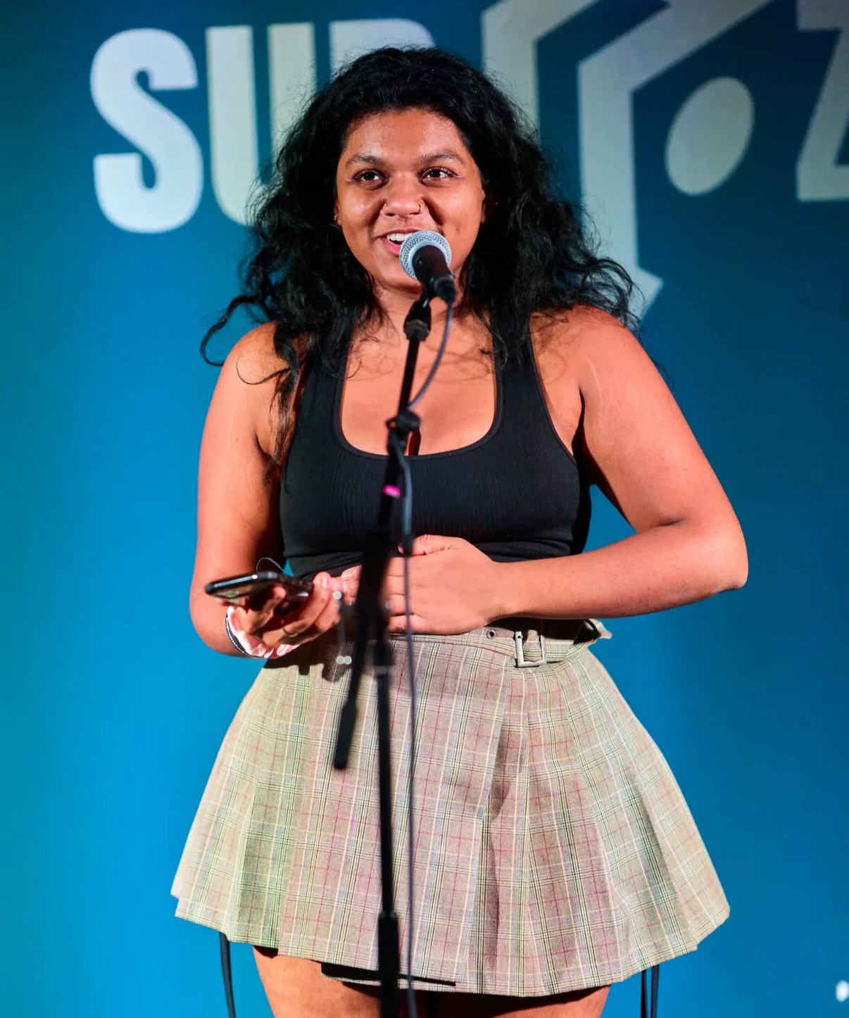 A woman with curly black hair is speaking into a microphone on stage, holding a phone in her left hand, and wearing a black tank top and a beige plaid skirt.