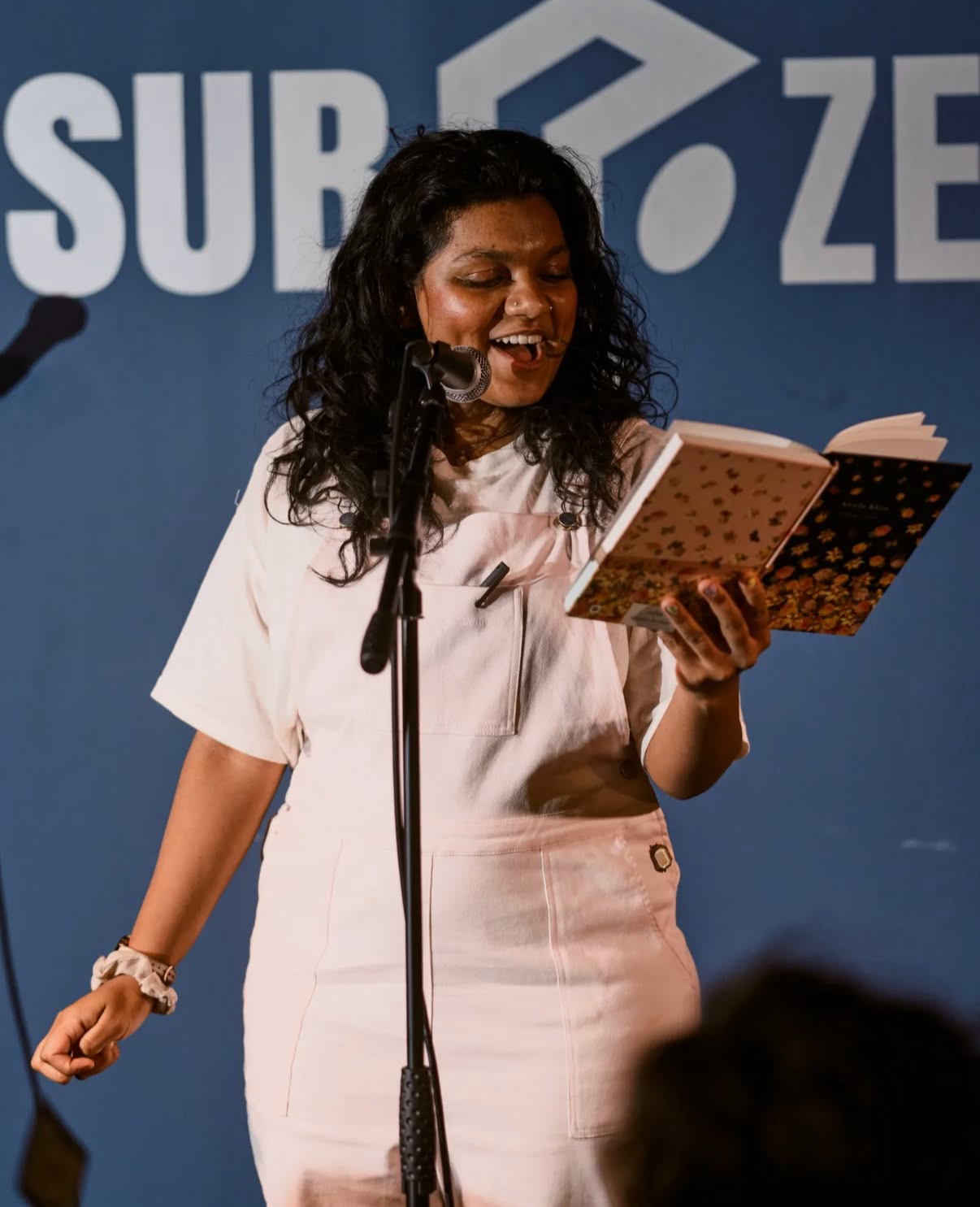 A woman with black curly hair, wearing a light pink outfit, is reading from a book at a microphone on stage with a blue background featuring the partially visible text 'SUBZ.'