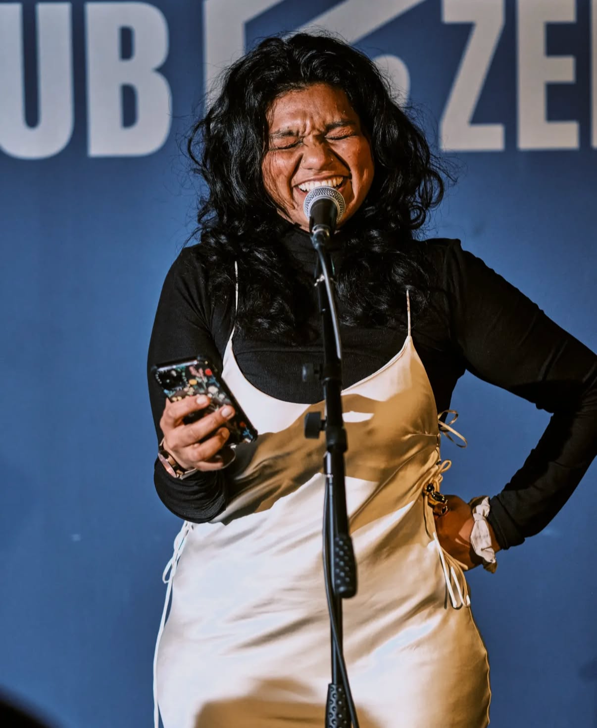 A woman with long black curly hair smiling and singing into a microphone while holding a phone in her right hand. She is wearing a black long sleeve shirt and a tan apron, standing in front of a blue background with white text.