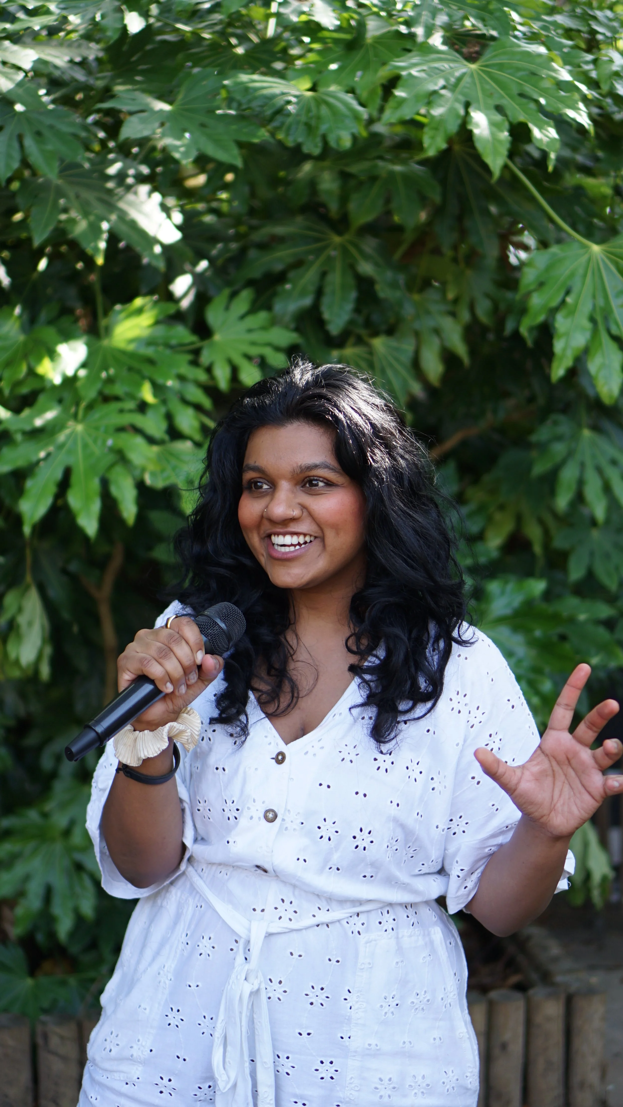 A NB femme with curly black hair holding a microphone and gesturing with her right hand while speaking outdoors in front of green leafy plants. Smiley, positive vibes.