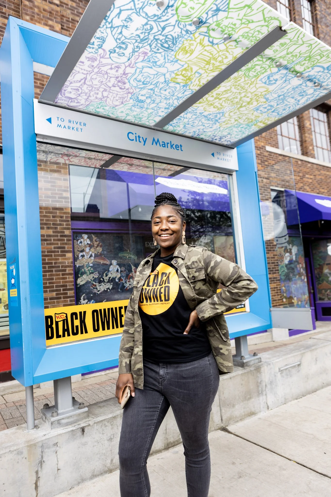 A young woman with braided hair smiling and standing in front of a vibrant bus stop shelter with a colorful mural on the roof, a sign indicating direction to River Market, and a yellow and black sign that reads 'Black Owned.' She is wearing a camouflage jacket, a black T-shirt with yellow lettering, and dark jeans, and holding a smartphone in her left hand.