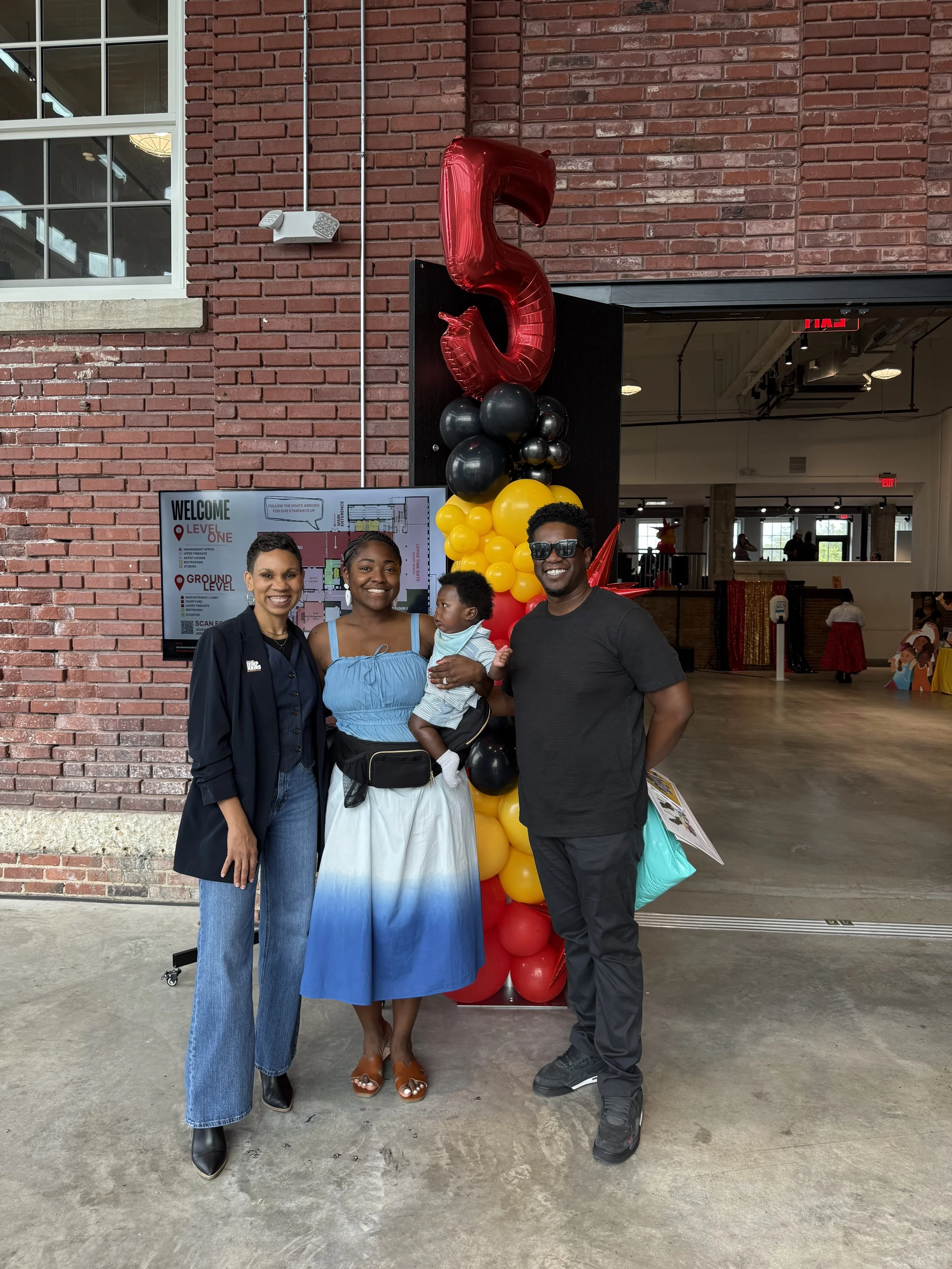 A group of four people standing in front of a brick wall decorated with black, yellow, and red balloons, featuring a large red number 5 balloon. They are smiling, and there is an indoor space visible behind them with several people and a table with a red and gold cloth in the background.