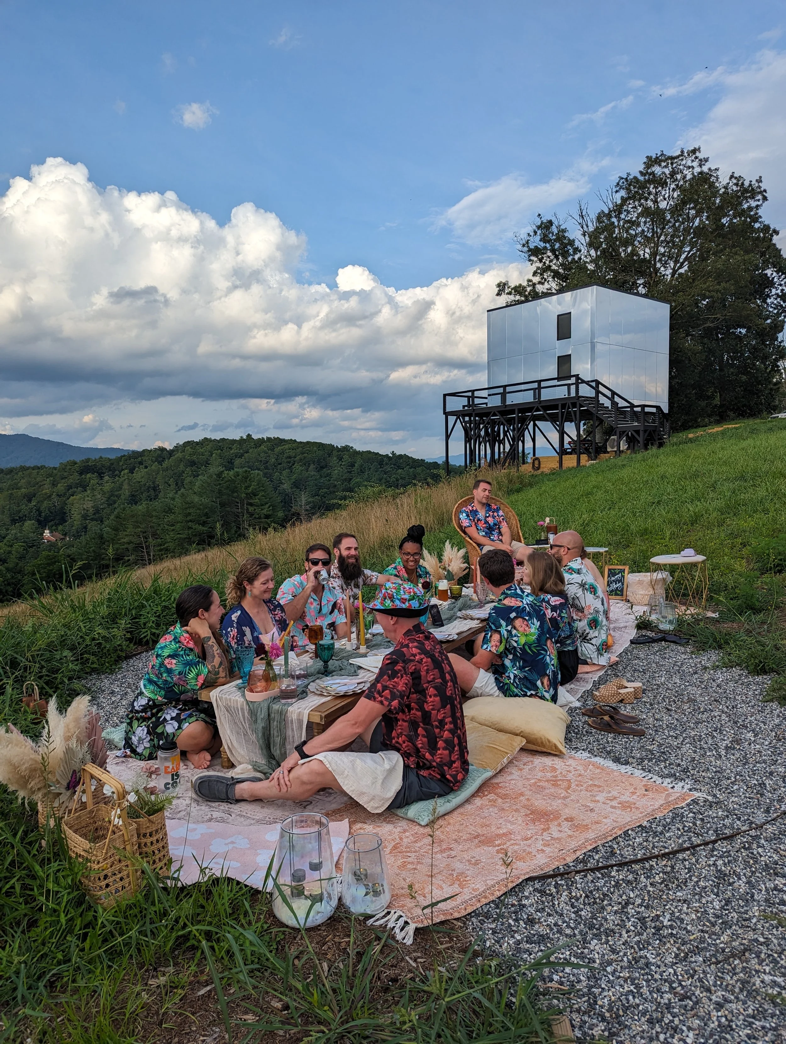 Luxury group picnic setup overlooking the Blue Ridge Mountains in Asheville, with guests seated around a styled low table featuring layered textiles, candles, florals, and curated tableware.