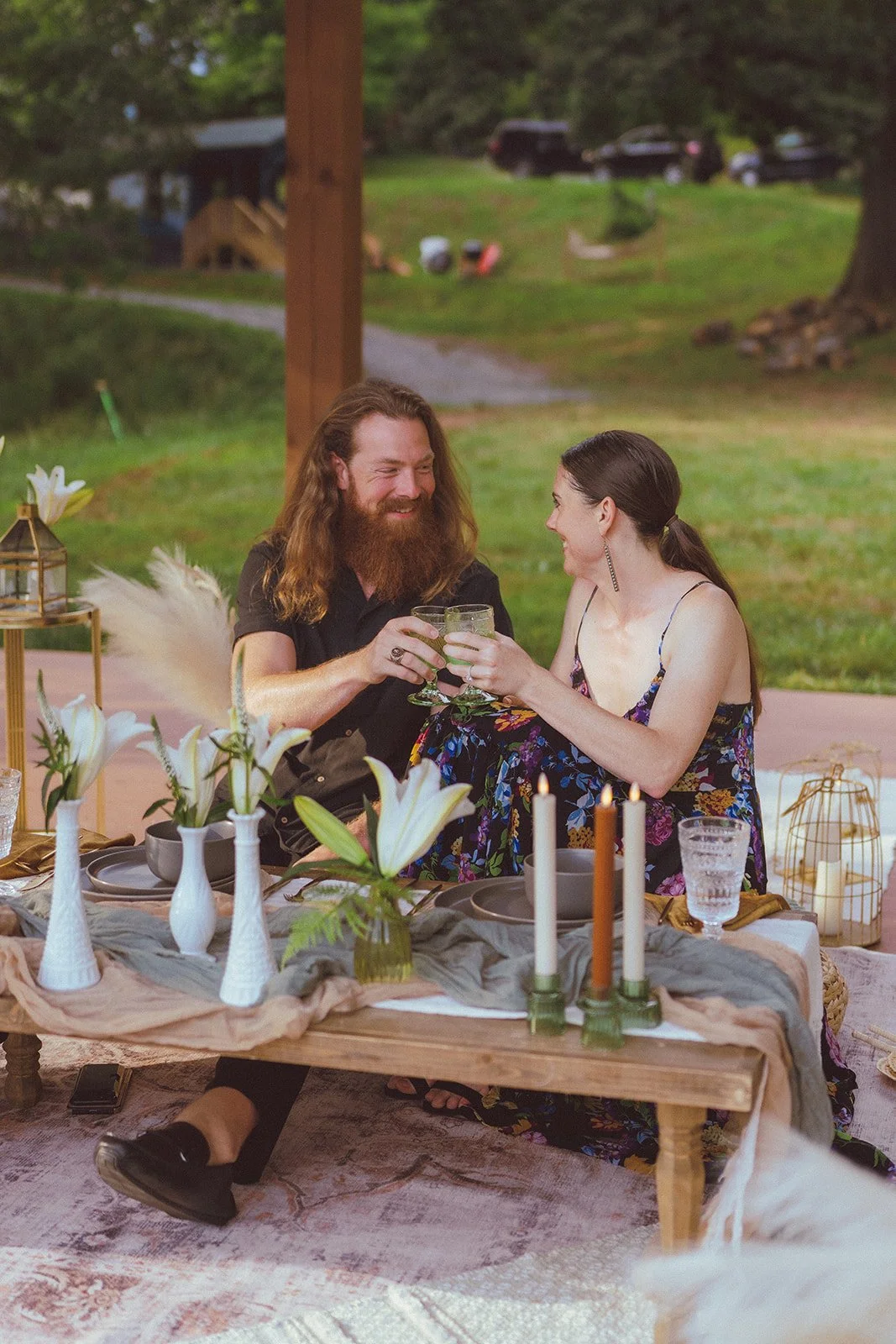 Couple toasting drinks at an intimate luxury picnic setup in Asheville, surrounded by candlelight, soft textiles, and fresh floral arrangements.