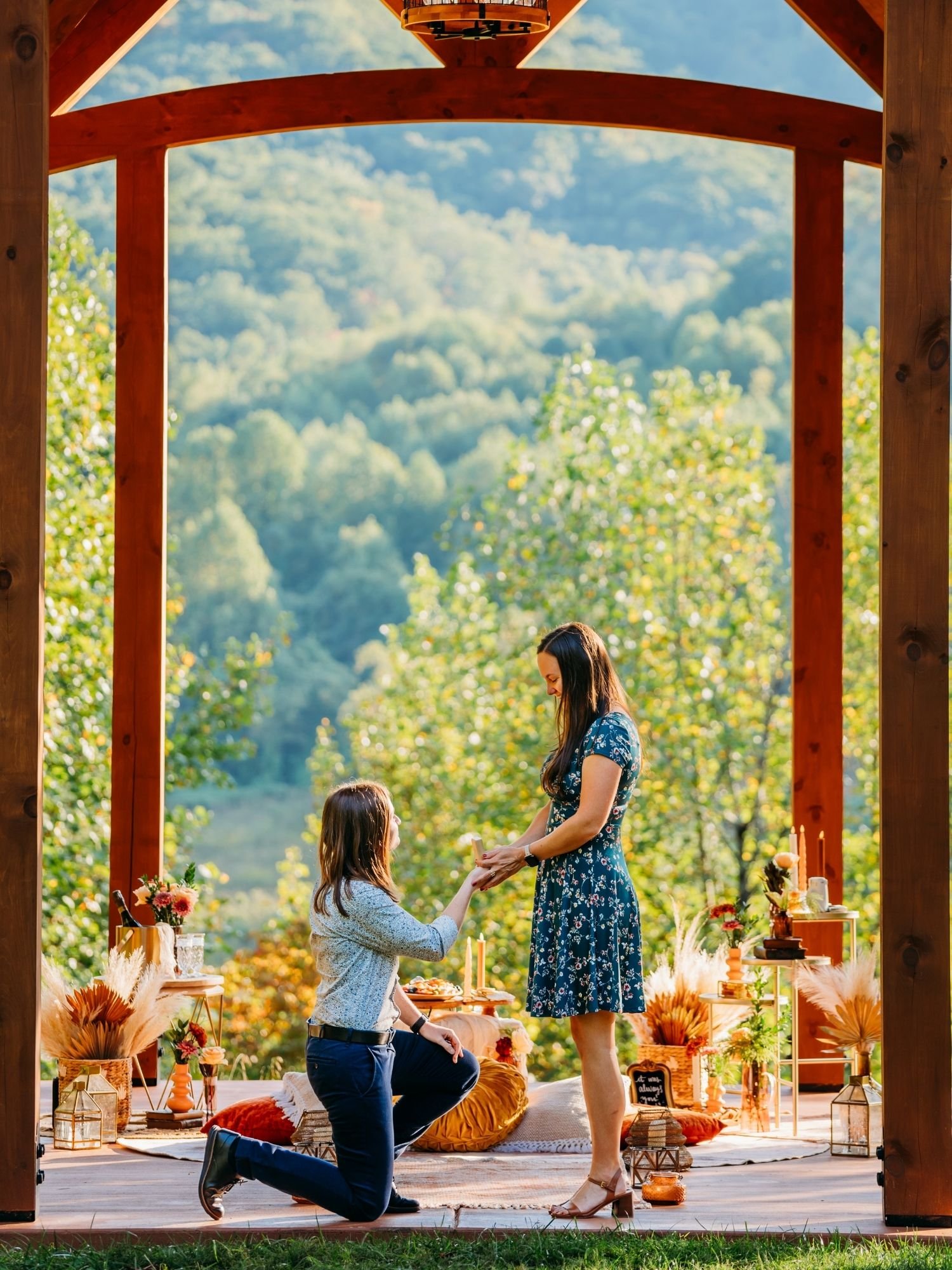 Surprise proposal picnic in Asheville with partner kneeling at a scenic mountain overlook surrounded by romantic decor and rose petals.