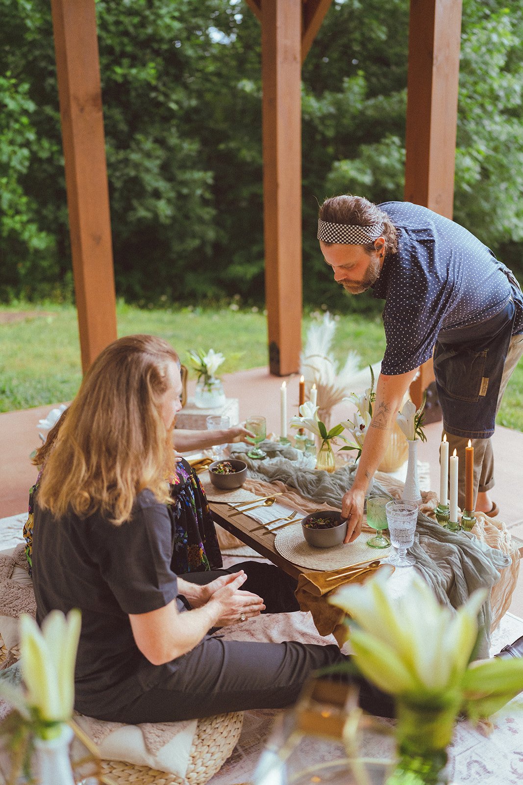 Host serving a beautifully styled luxury picnic table with layered linens, gold flatware, candles, and fresh florals at an outdoor venue in Asheville.