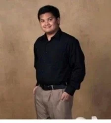 A young man in a black shirt and beige pants standing against a beige background, smiling at the camera.