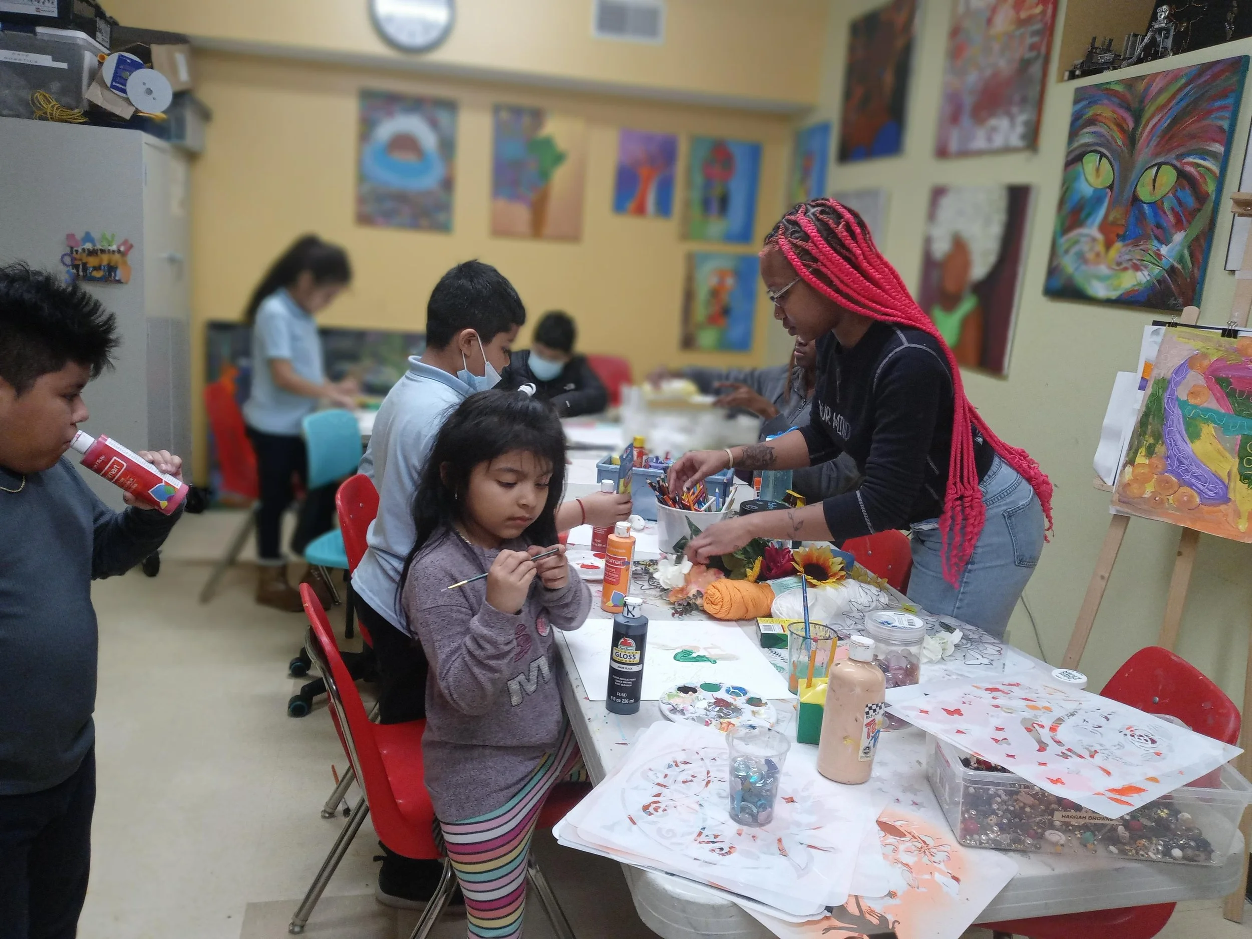 Children and an adult participating in an art class with various paintings on the walls and art supplies on the table.