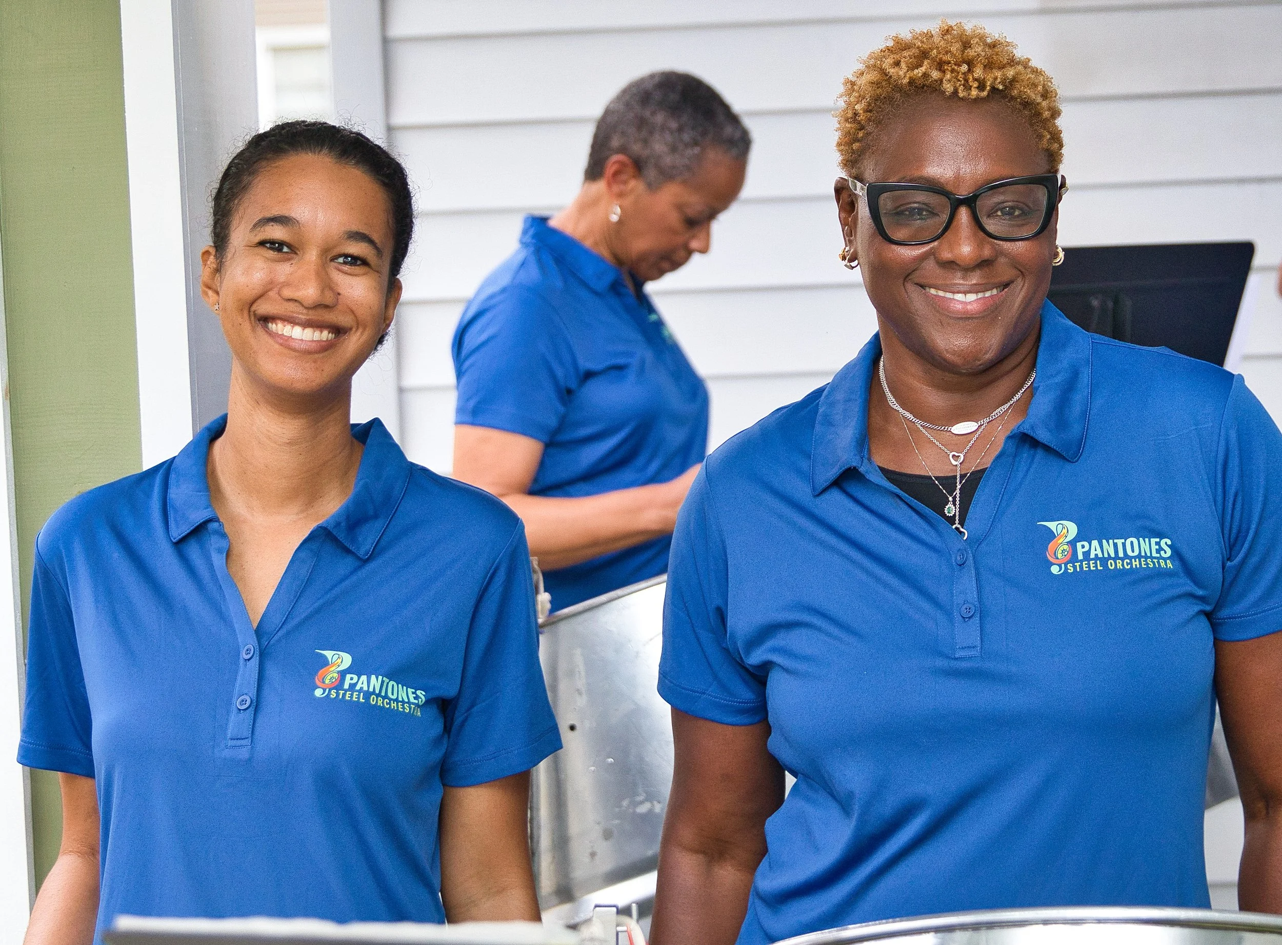 Three women wearing matching blue shirts with the Pantones Steel Orchestra logo, smiling outside.
