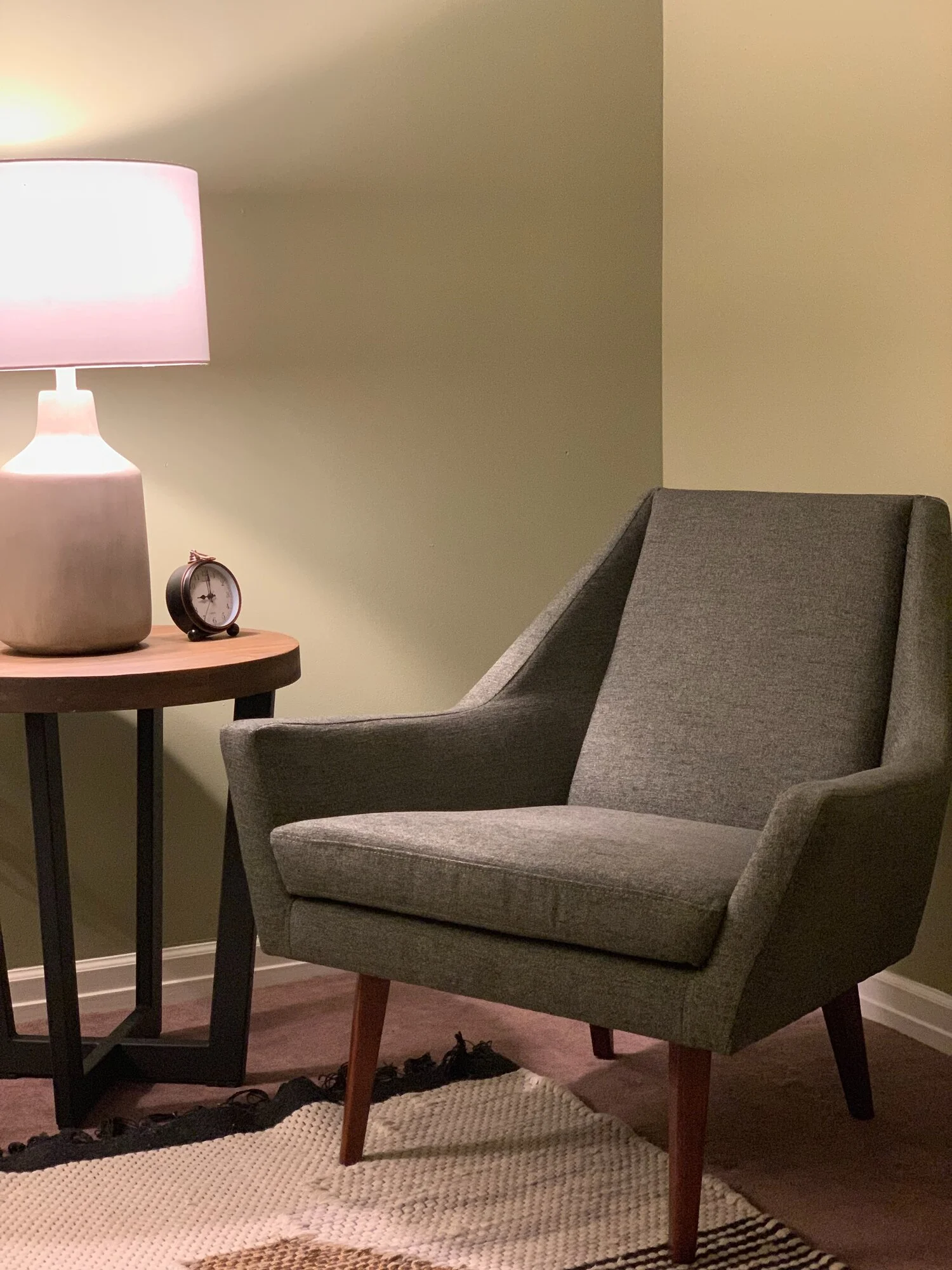 A cozy corner with a gray armchair, a wooden side table holding a large lamp with a pink shade, and a small round clock. There is a patterned rug on the floor and the background is a two-tone wall.
