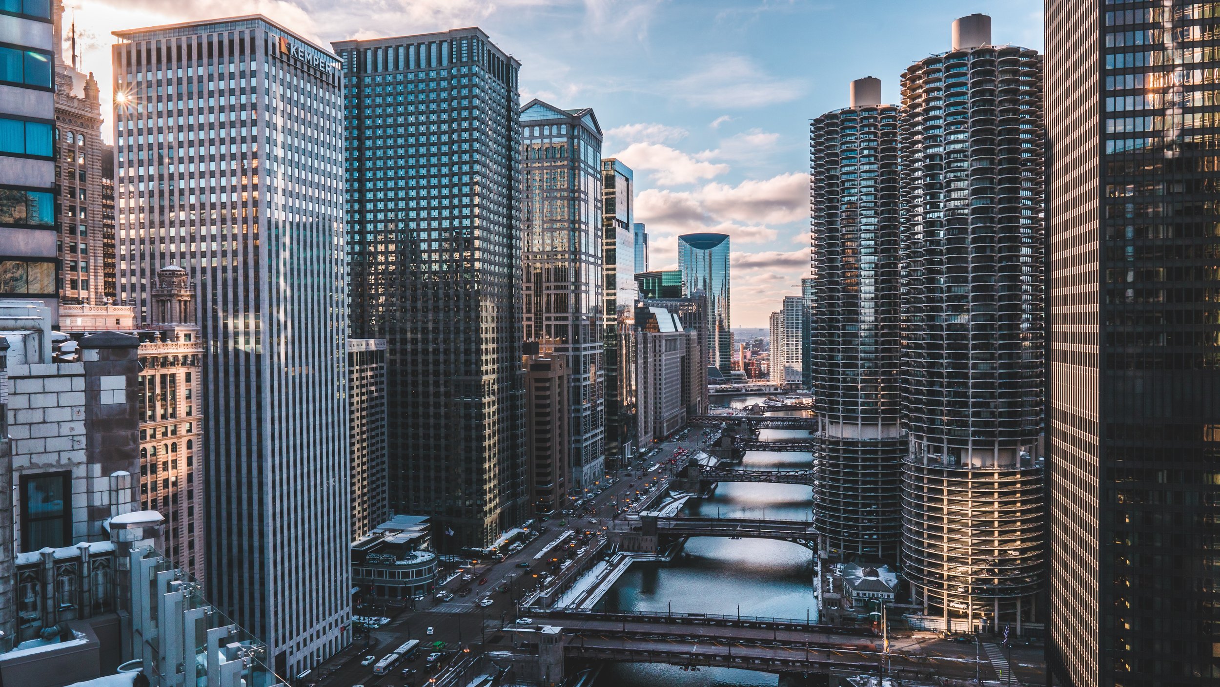 A cityscape of downtown Chicago featuring tall skyscrapers lining the Chicago River, with bridges crossing over the water and a partly cloudy sky overhead.