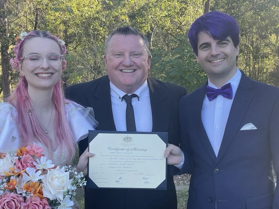 Three people standing outdoors, smiling, with a marriage certificate. The person on the left is a woman with pink hair and glasses, holding a bouquet of flowers. The person in the middle is a man in a black suit, white shirt, and black tie, holding the marriage certificate. The person on the right is a man with dark purple hair, wearing a tuxedo with a purple bow tie and pocket square.