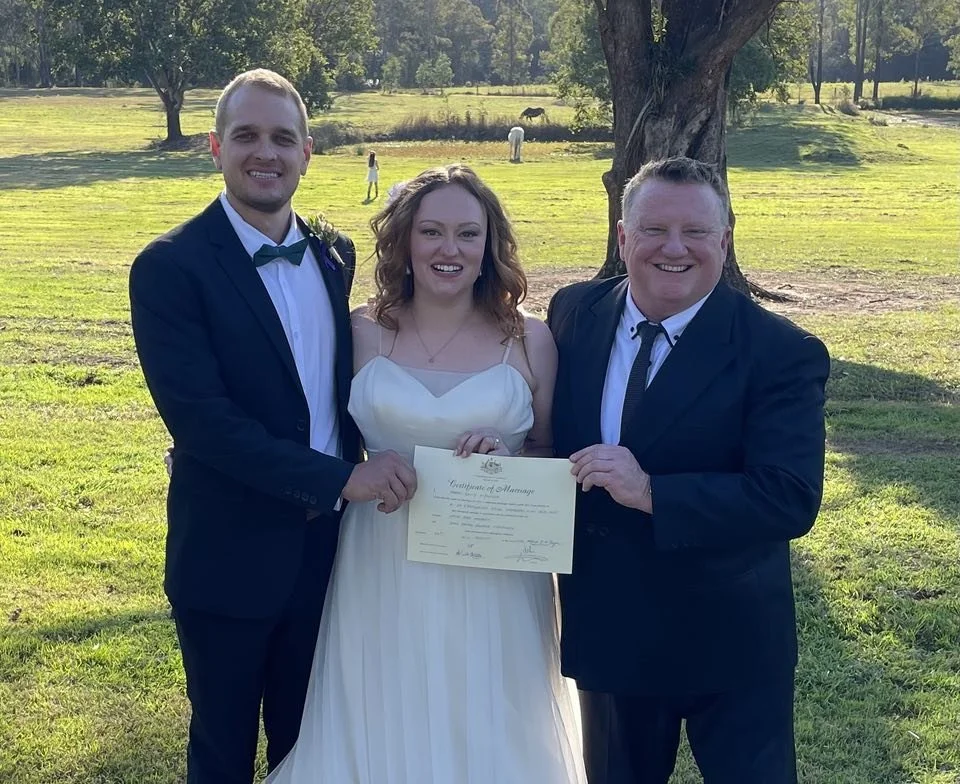 Two men and a woman in wedding attire smiling outdoors, holding a marriage certificate, with trees and a grassy field in the background.