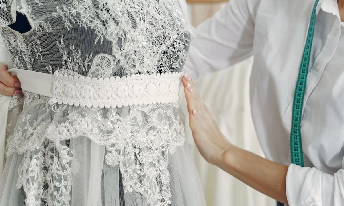 Custom wedding dress fitting showing floral lace fabric, decorative bridal waist detail and a designer measuring the gown