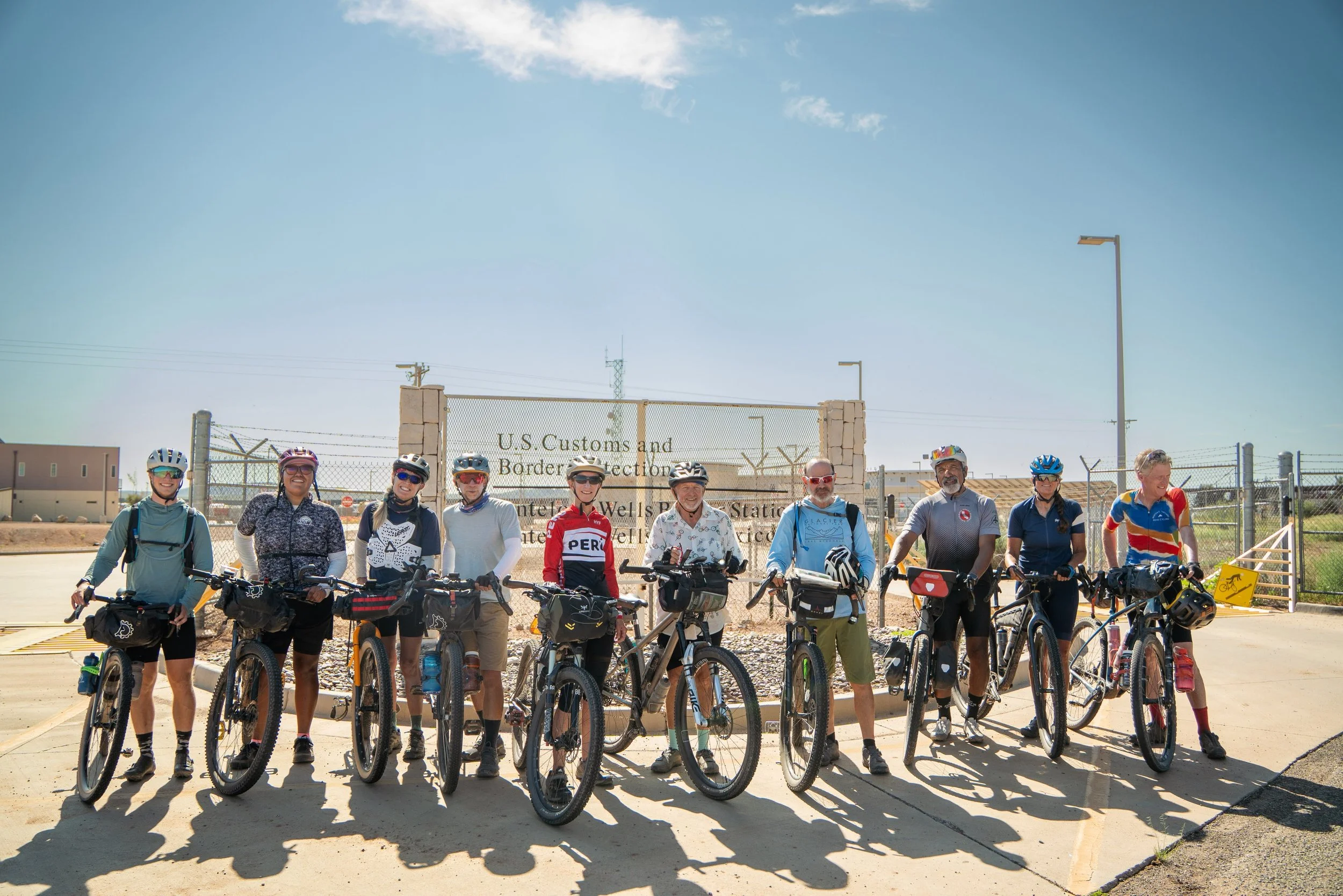 Ten cyclists standing with their bikes at the Antelope Wells border crossing sign in New Mexico, the southern end of the Great Divide Mountain Bike Route