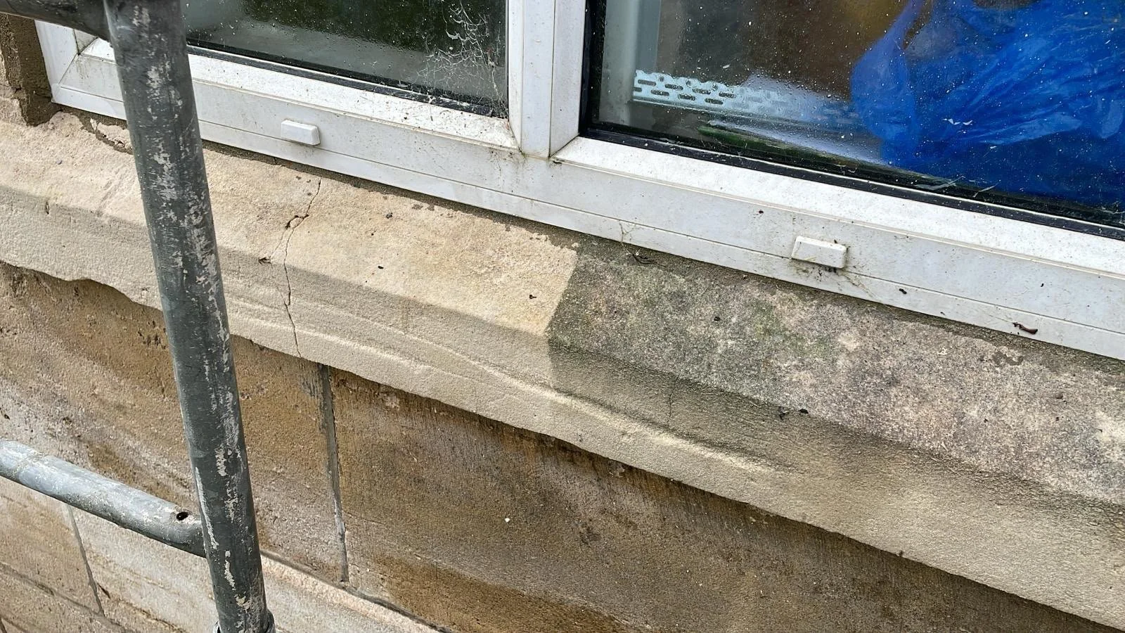 Close-up of a house window with a white frame and sills, cracked stone brick wall, and a metal pipe in front.