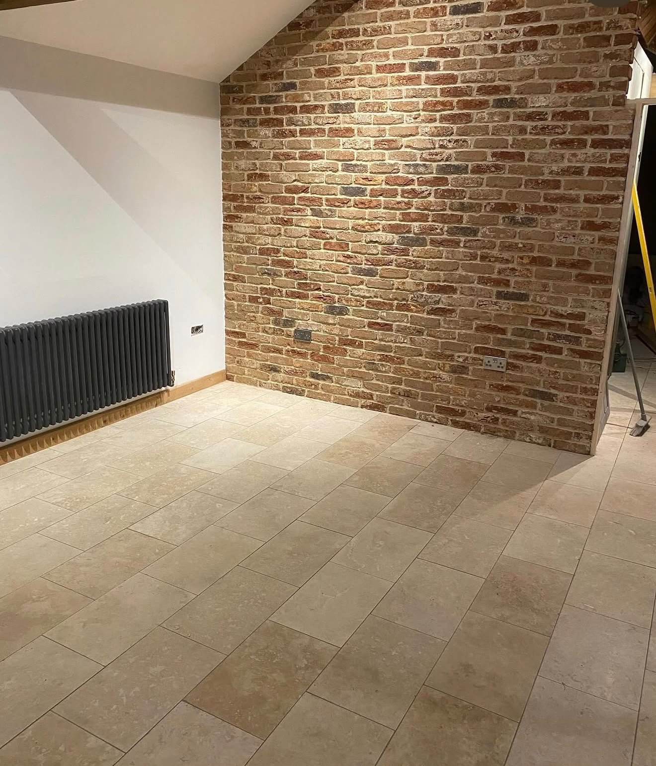 Interior of a room with beige tiled flooring, a red brick accent wall, a white wall with a black radiator, and multiple electrical outlets.