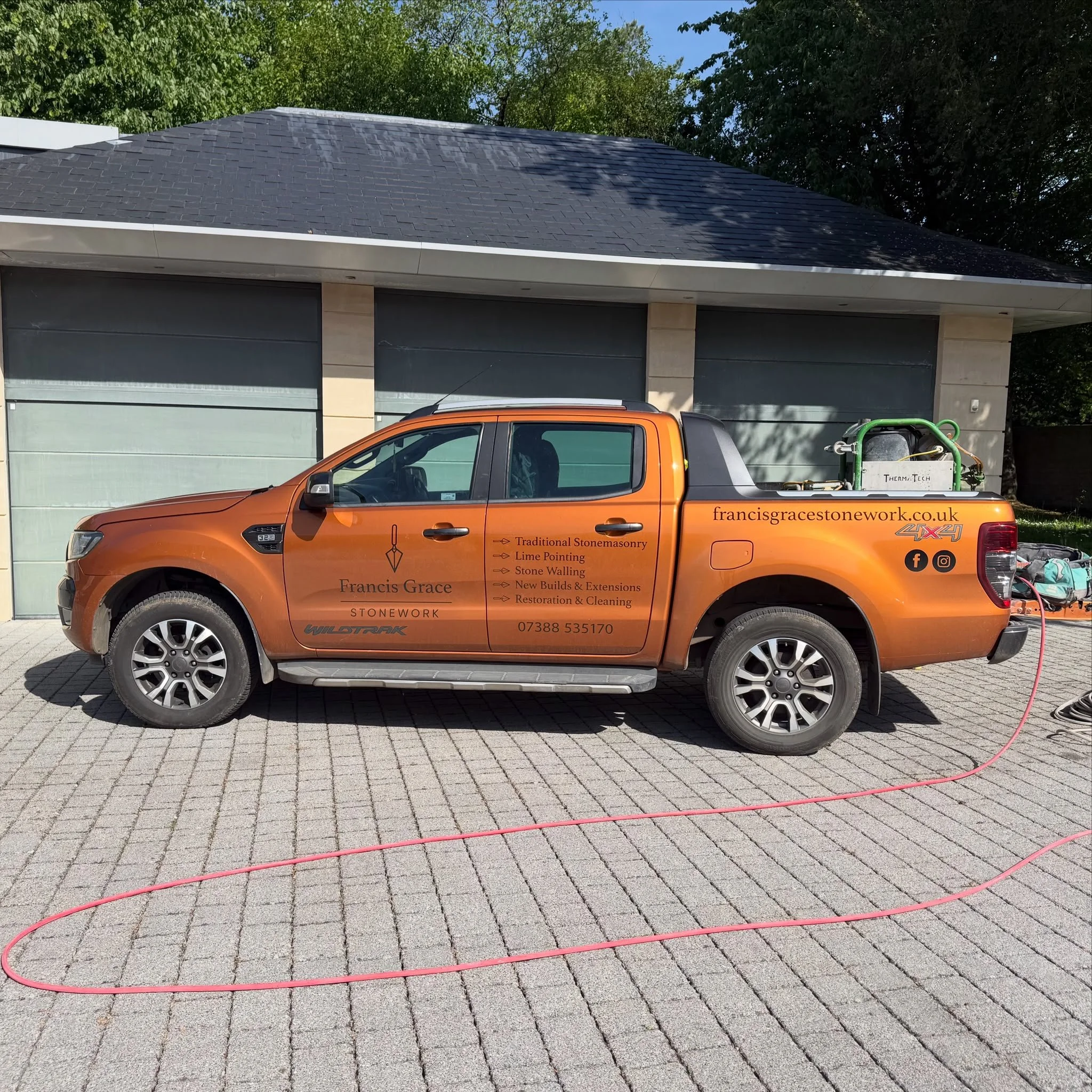 Construction worker truck parked on a paved driveway in front of a garage with closed doors, orange truck with signage for stonework services, ropes laid out on the ground, green bushes and trees nearby.