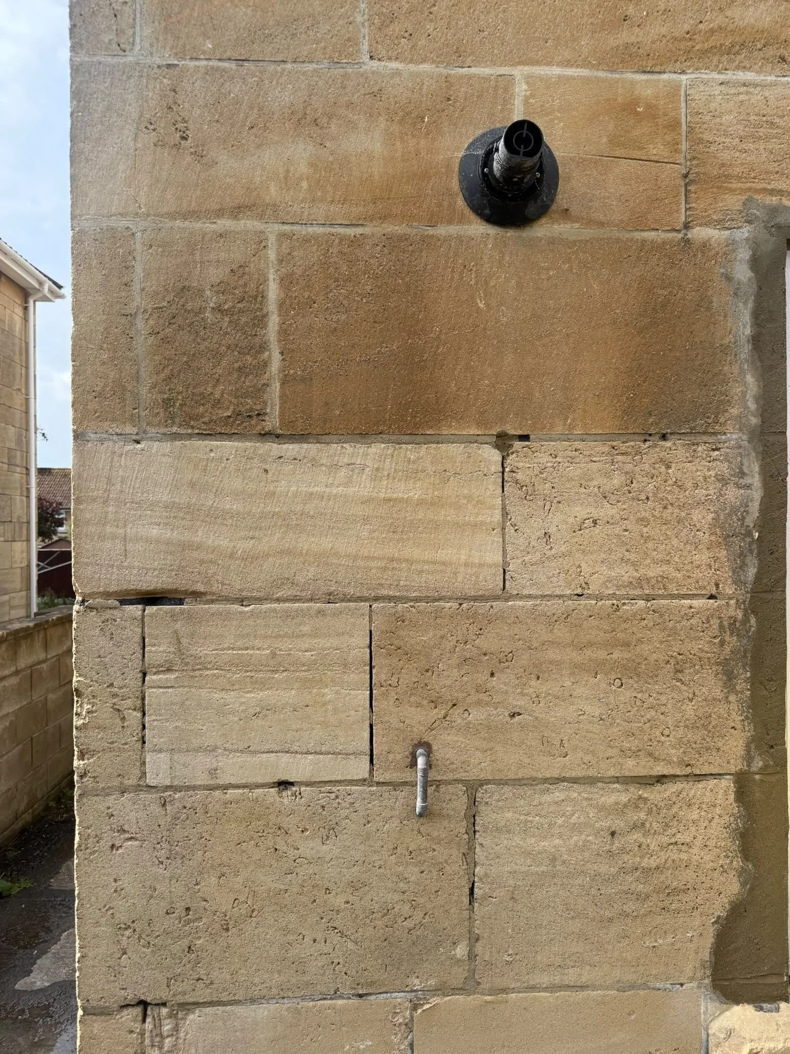 Close-up of a sandstone wall with a black circular fixture and a small pipe protruding from the wall.