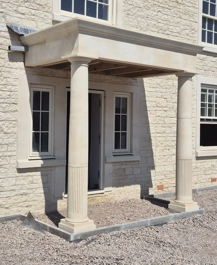 View of a house with a small porch supported by two classical columns, on a gravel ground with a brick border.