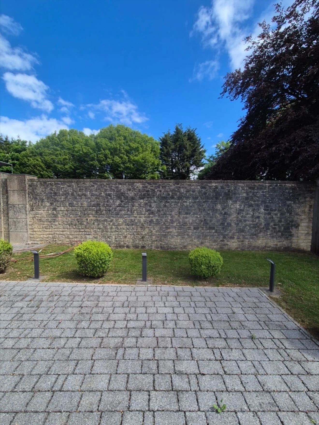 A paved courtyard with small green bushes, a stone brick wall, and trees in the background under a partly cloudy blue sky.