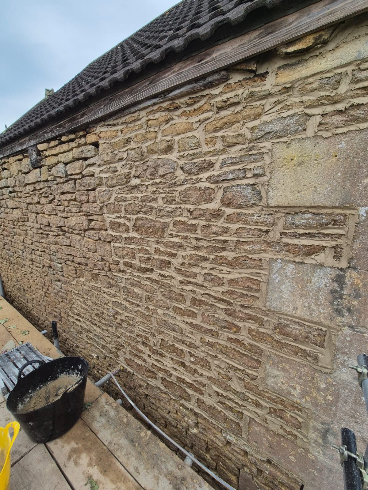 A stone brick wall under construction with a wooden beam and tiled roof above. Construction tools and materials are placed at the ground level.