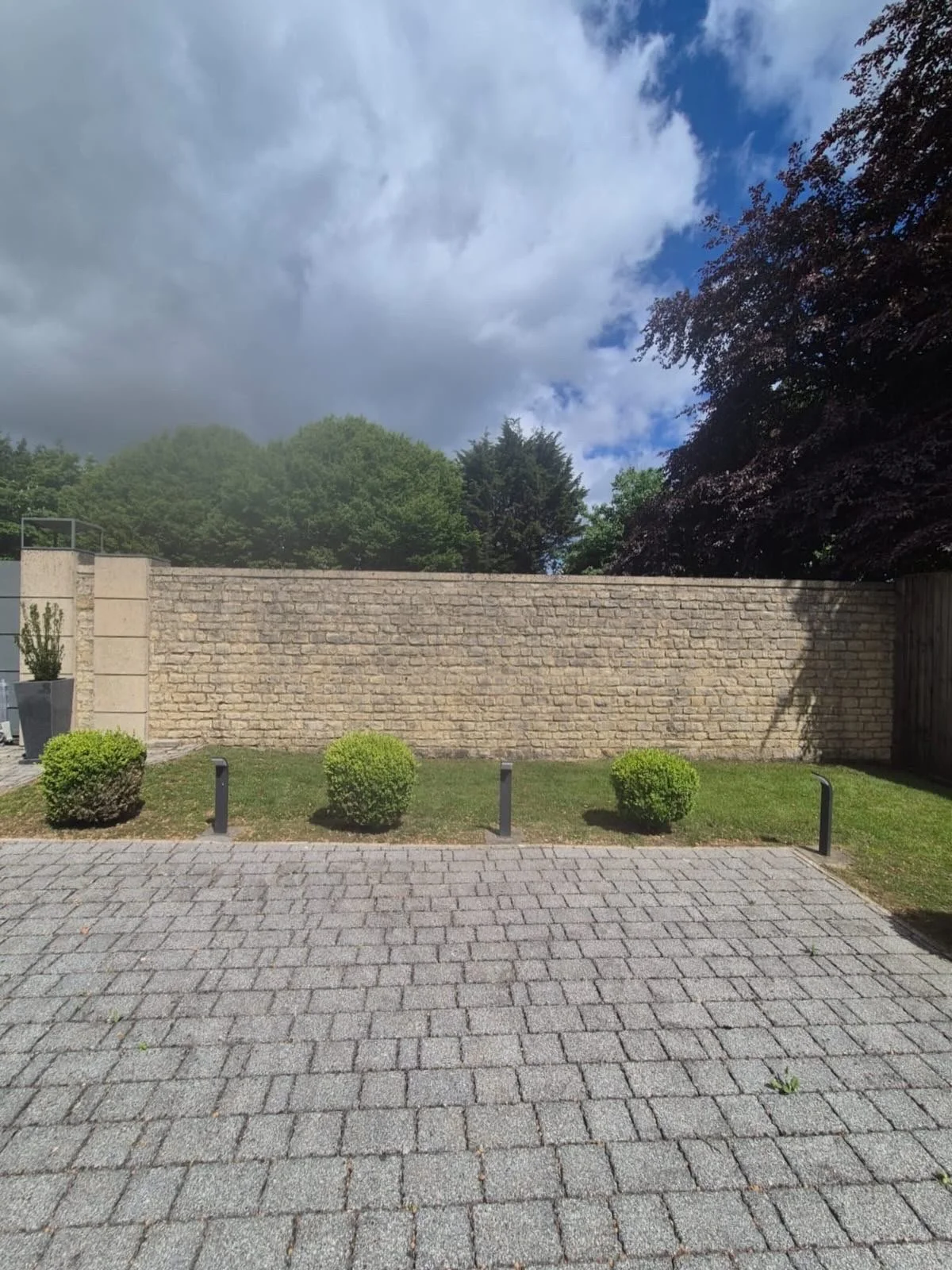 A paved brick courtyard with a low brick wall, small bushes, and tall trees in the background under a partly cloudy sky.