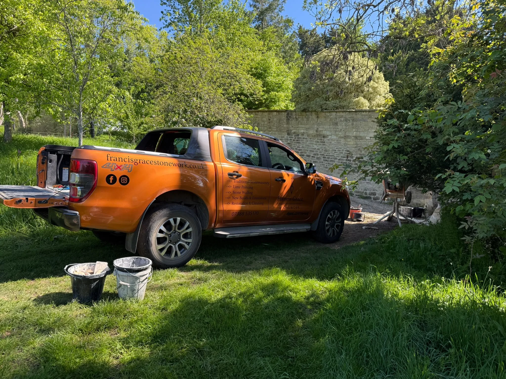 An orange pickup truck with company branding parked on a grassy area with trees and a brick wall in the background.