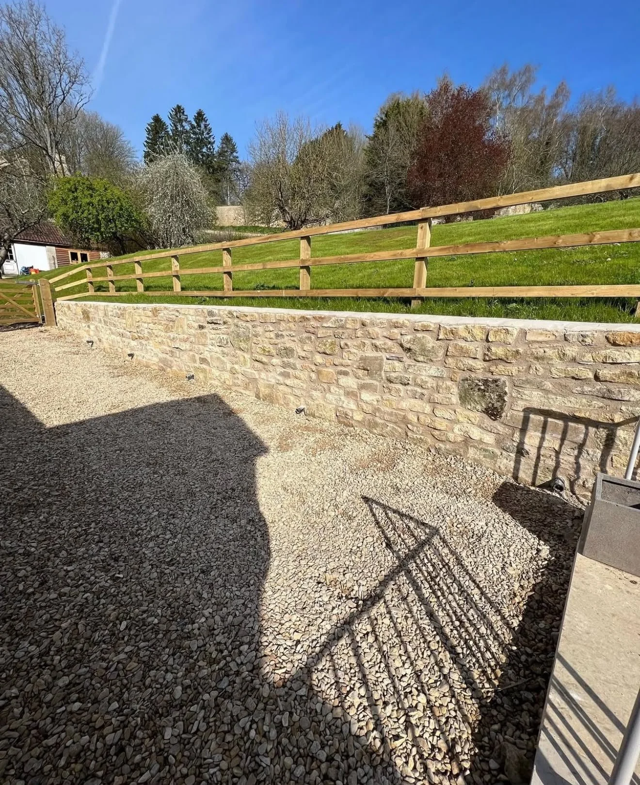A gravel patio area in front of a stone retaining wall, with a shadow of a chair cast on the gravel. A wooden fence separates the patio from a grassy hill with trees in the background and a clear blue sky.