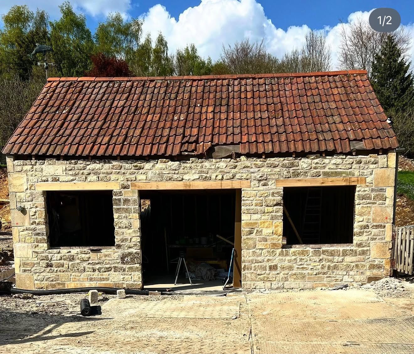 A small brick building under construction with three large window openings and a doorway, a pitched red tile roof with some damage, surrounded by construction materials, and trees in the background.