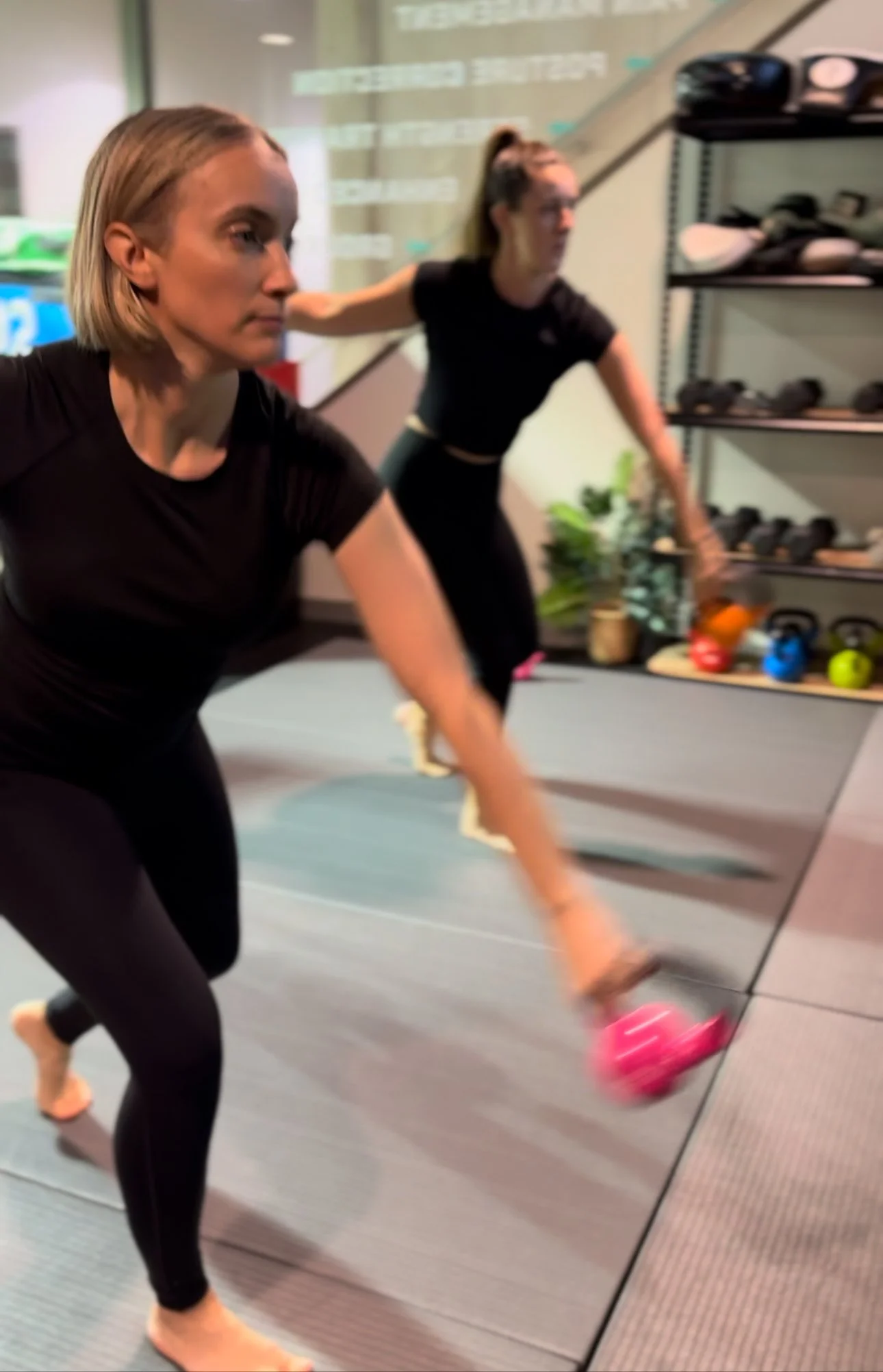 Two women exercising with dumbbells in a gym, one in the foreground lifting a pink dumbbell and the other in the background reaching towards the floor.