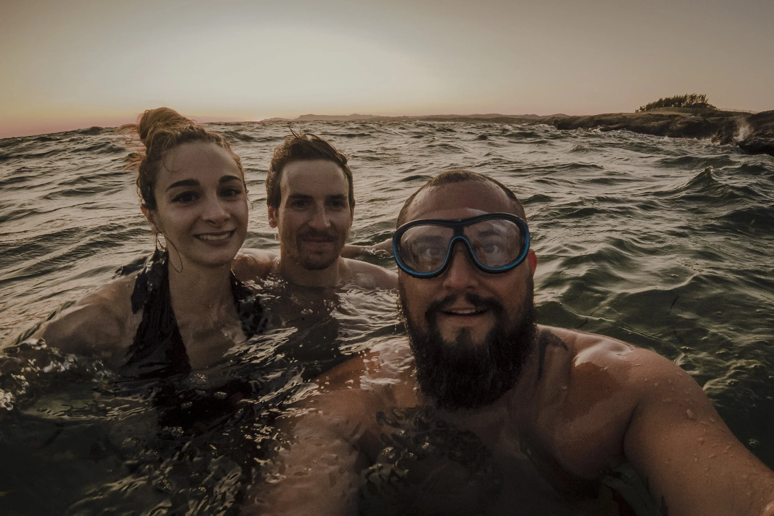 Trois personnes prennent un selfie dans la mer au coucher du soleil, un homme avec des lunettes de plongée, une femme avec les cheveux relevés, un autre homme à l'arrière