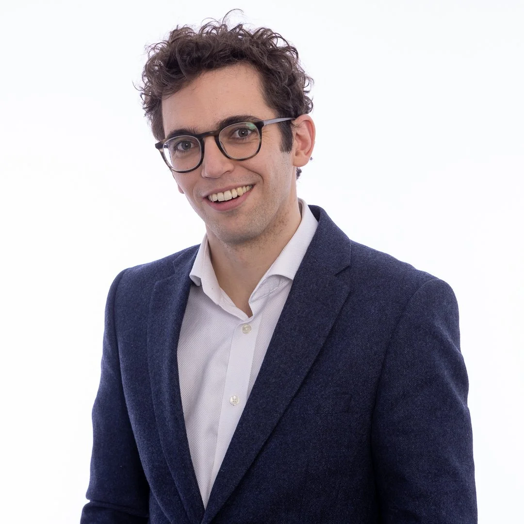 Professional young man with curly brown hair and glasses, smiling in a business suit against a white background.