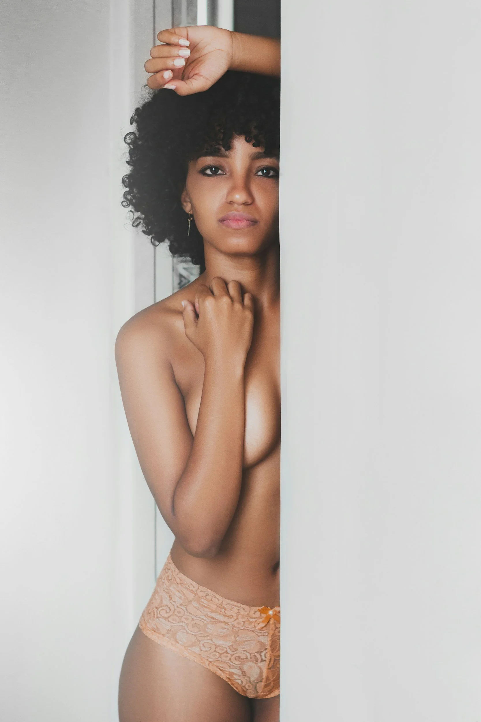 Mulher de pele morena com cabelo cacheado, escondida parcialmente atrás de uma parede branca, usando lingerie de calcinha de renda em tom pêssego. Ela olha para a câmera com expressão séria, com uma mão tocando o pescoço e a outra levantada, apoiada na parede.