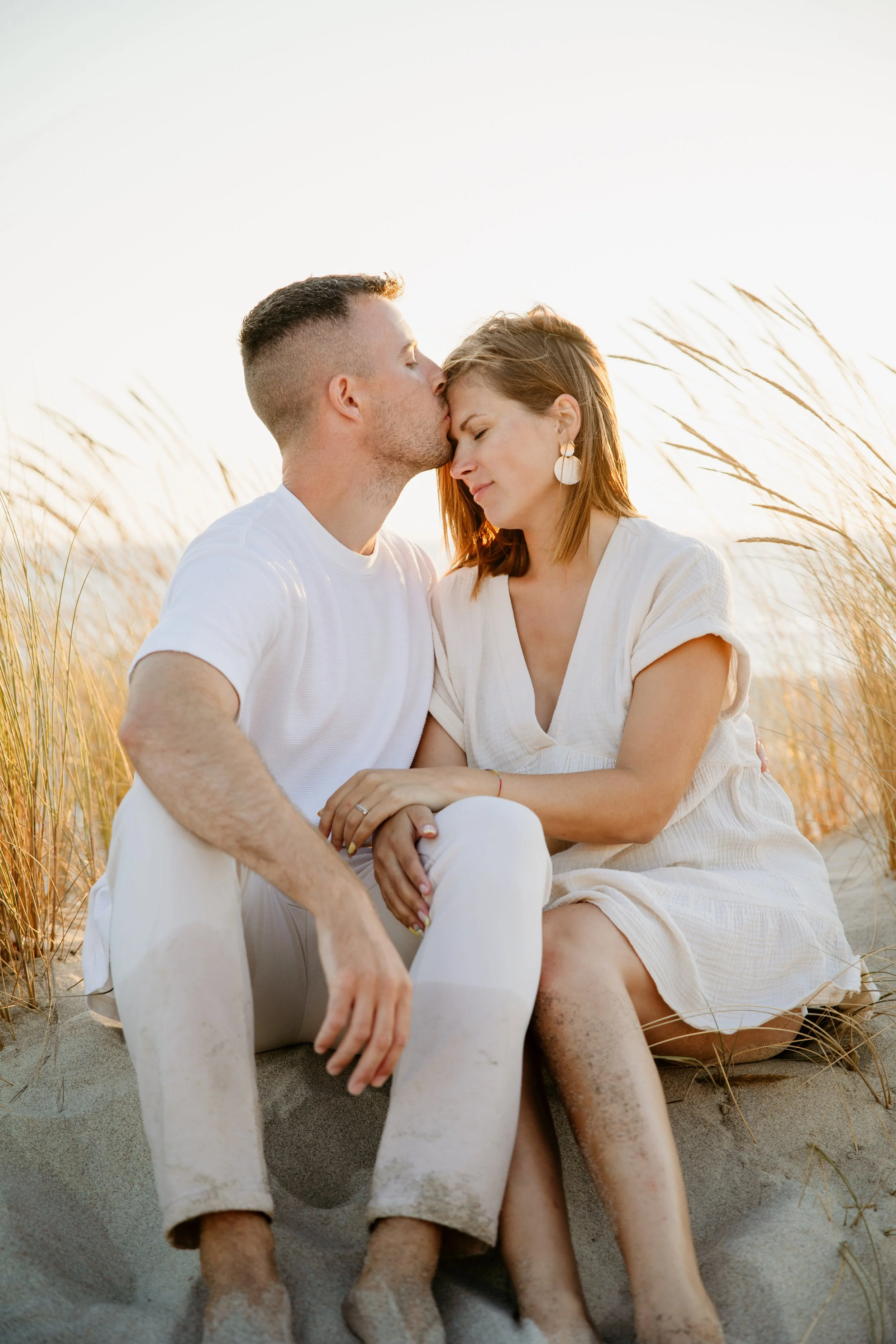 Casal na praia, com o homem beijando a testa da mulher, que sorri com os olhos fechados, ambos sentados na areia entre gramíneas ao entardecer.