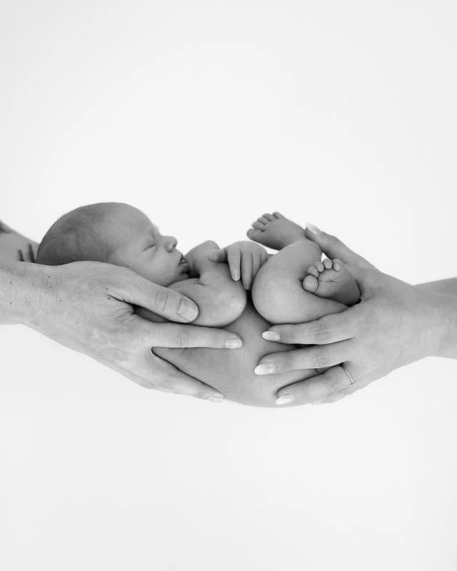 Bebê recém-nascido deitado em uma manjedoura, cercado por mãos humanas que o sustentam com carinho.