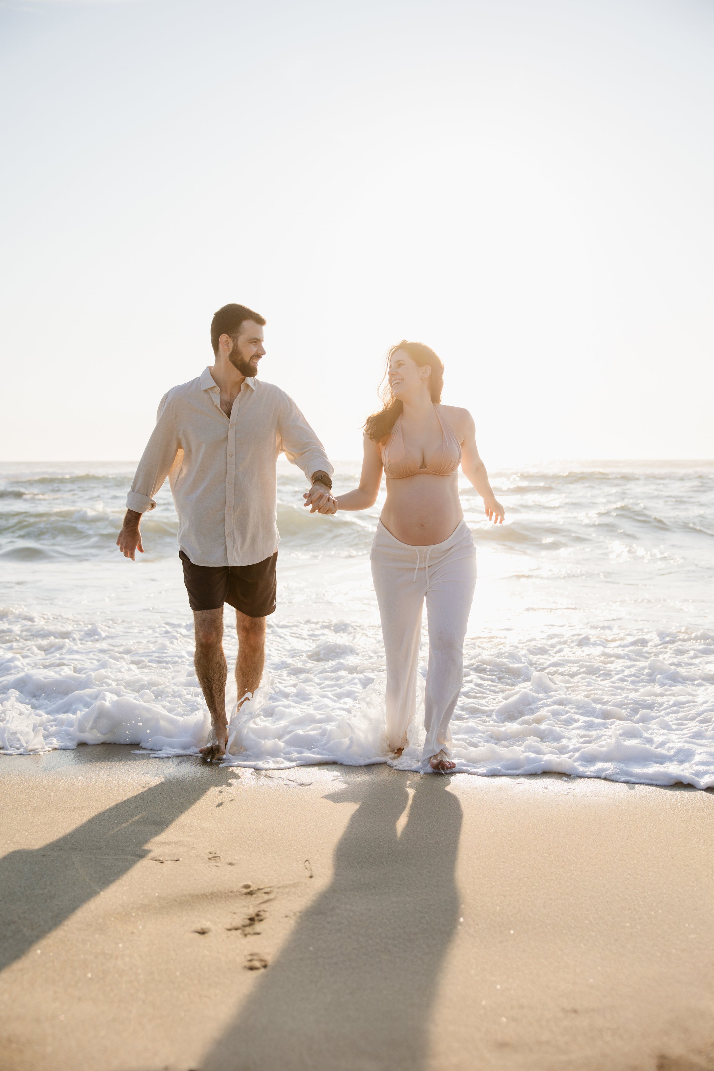 Casal feliz caminhando na praia ao pôr do sol, com o mar ao fundo, mostrando momentos românticos e de lazer.