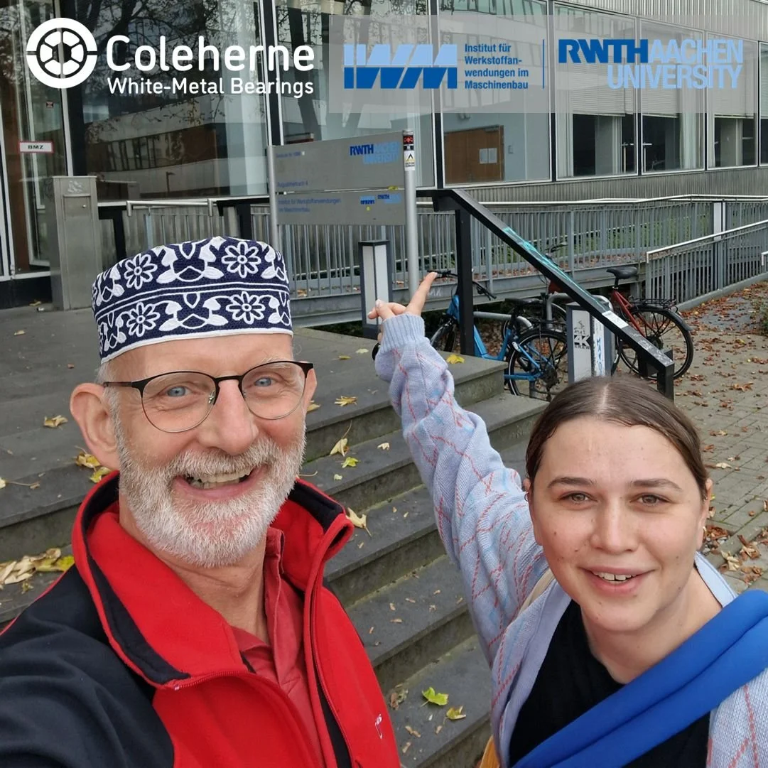 Two people, an older man with glasses wearing a patterned hat and a red jacket, and a young woman in a light hoodie and black shirt, smiling and taking a selfie outside a university building with bicycles nearby. The building has signage for RWTH Aachen University and the Institute for Material Science.